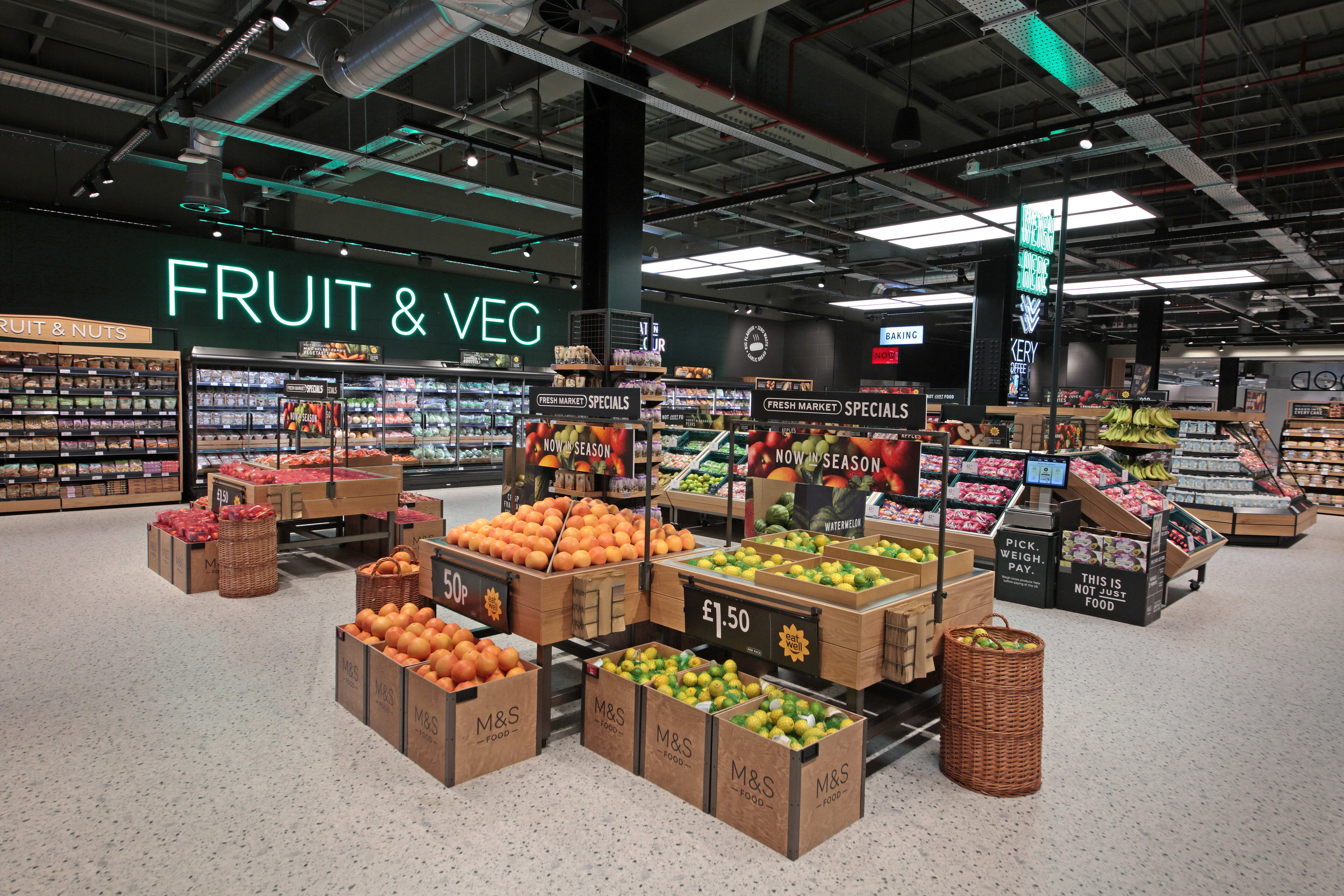 Marks & Spencer supermarket fruit and veg section with vivid lighting. Displays of oranges and apples in wooden crates, labels highlight prices and seasonal specials.