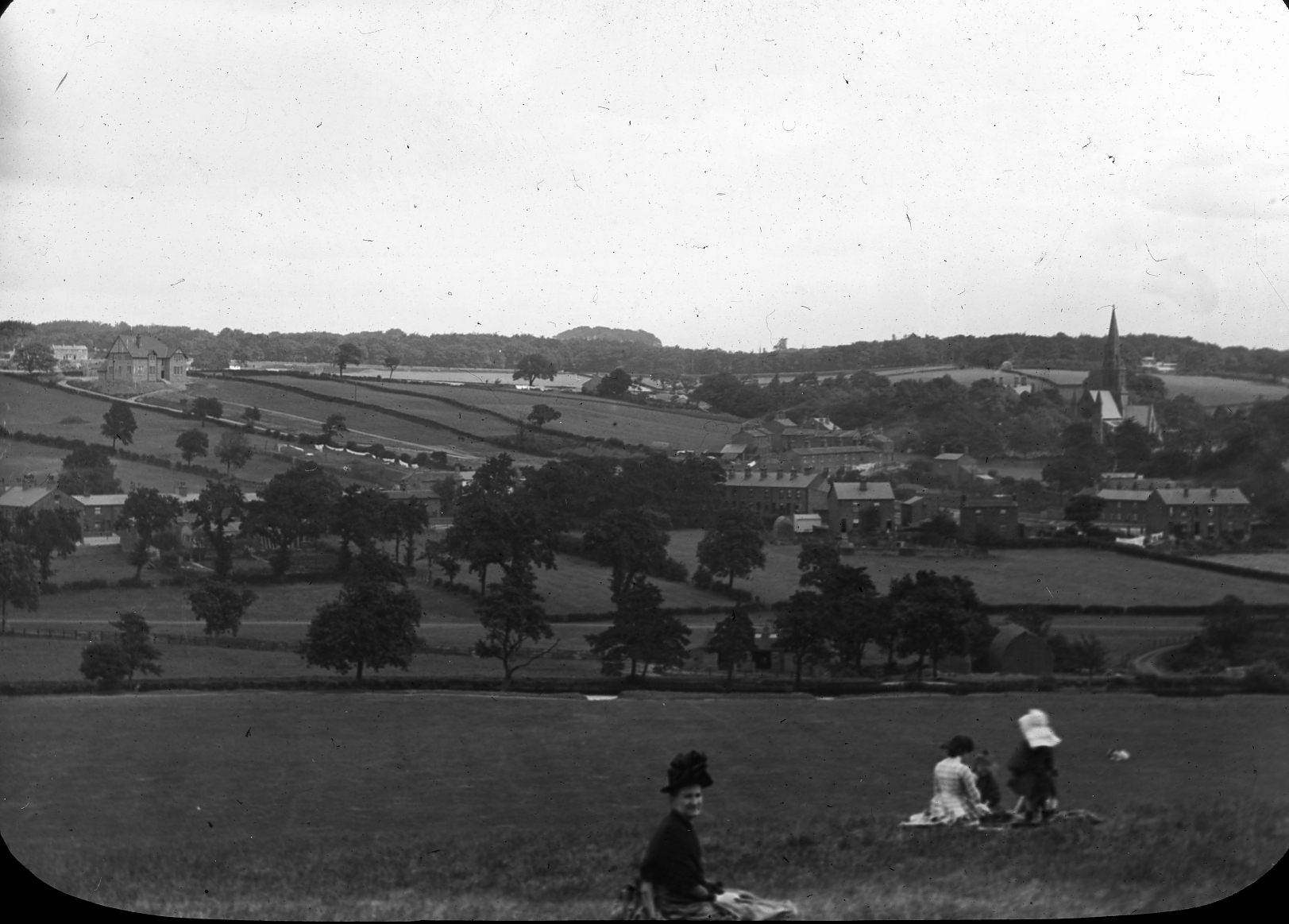 Black and white photograph of fields and trees. The land dips to a valley in front of the camera and then rises again to a hill. There is a church and houses in the middle distance on the right and a large house on the left. In the foreground sit two adult females and two children.