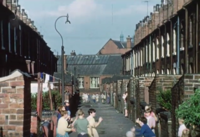 Colour film still showing children playing in a back alley, with terraced houses on each side.