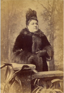 Black and white portrait of Mary Brewer.  She is wearing a fur coat and her hari is piled up on top of her head.  The backdrop is of painted trees.