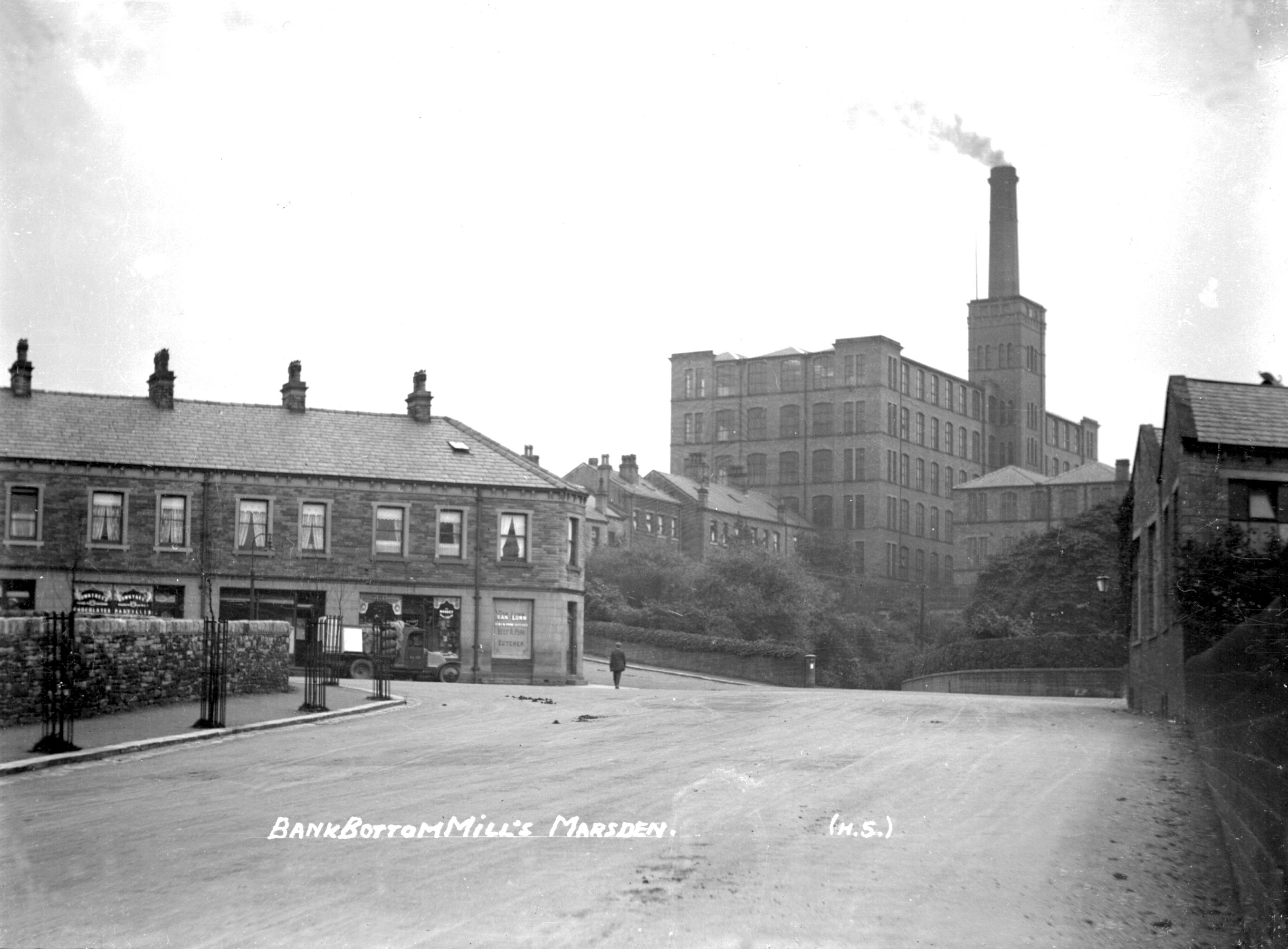 A historic black-and-white photograph showing Bank Bottom Mills in Marsden, with its name written in white text in the lower part of the image. The mill's tall smokestack emits smoke. Shops line the street, and one person walks along the otherwise empty road.