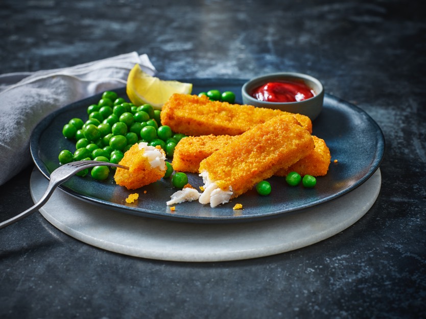 A dark plate with three golden fish sticks, bright green peas, a lemon wedge, and a small dish of red sauce, conveying a fresh and appetizing meal.