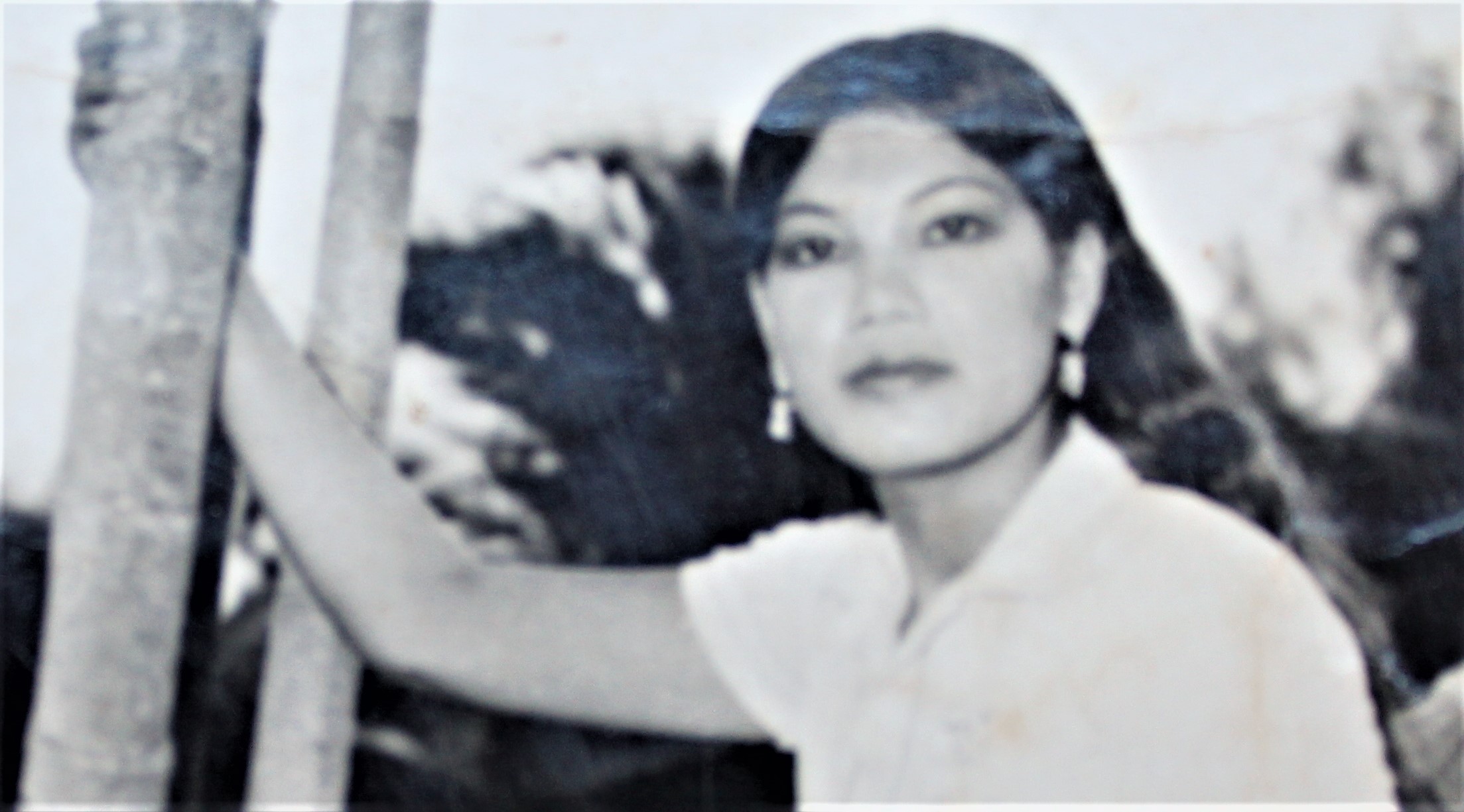 The image is a black-and-white photograph of a young woman named Sophari Ashley standing outdoors. She has long, dark hair that falls past her shoulders and is wearing earrings. Sophari Ashley is gazing directly at the camera with a calm expression. She is holding onto a tree with one hand, which adds a natural element to the scene. Her attire is simple, with a light-colored top. The background is slightly blurred, with some foliage visible, giving the image an outdoor, possibly tropical, setting. The photo has a vintage feel, suggesting it was taken several decades ago.