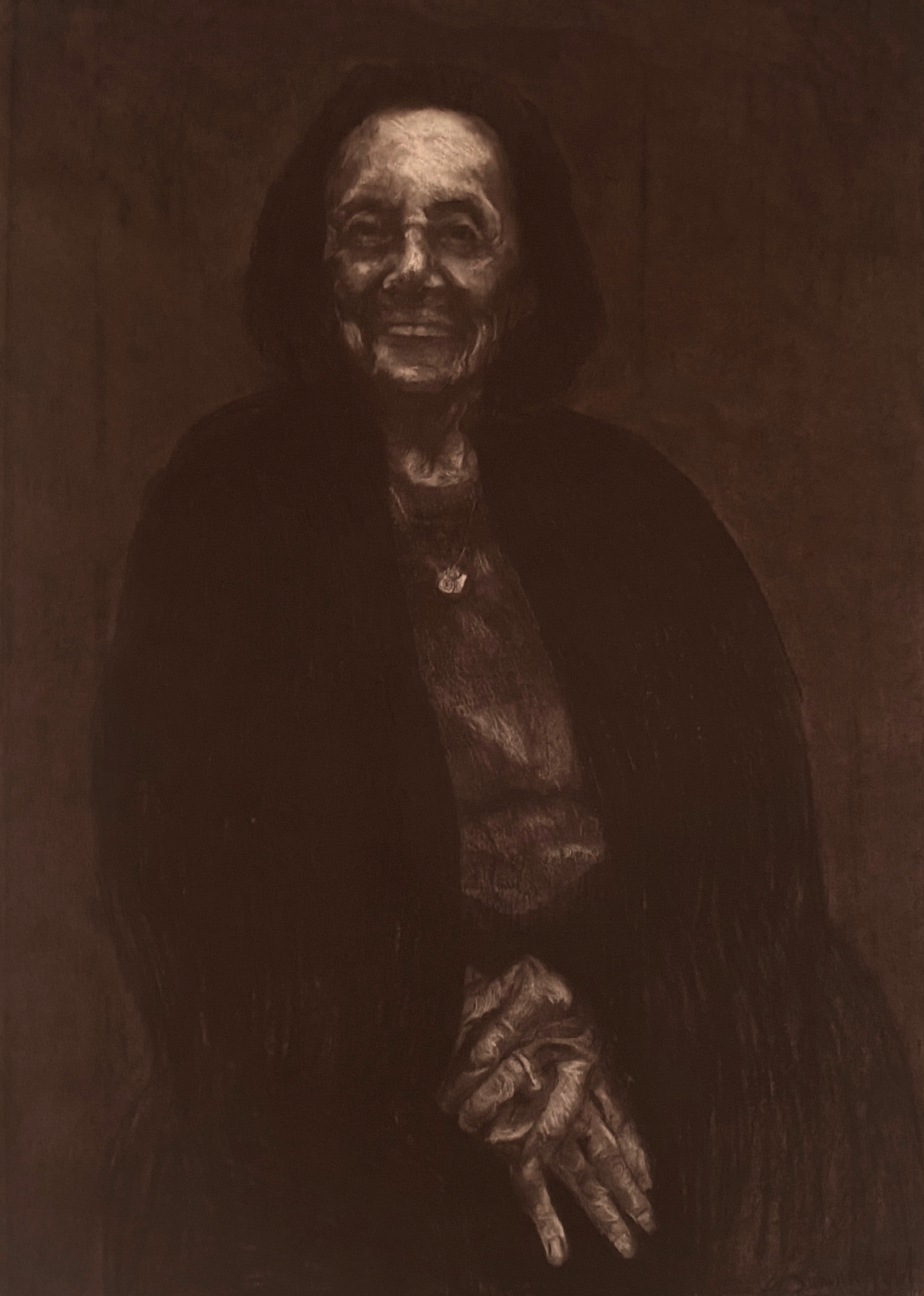 The image is a dark, monochromatic portrait of an elderly woman. The lighting is subdued, creating deep shadows that envelop much of her figure, leaving only parts of her face, hands, and upper body more clearly visible. The woman has a gentle smile, and her expression conveys warmth and resilience. She is wearing a dark garment, possibly a shawl, over a top, and a small pendant necklace is visible around her neck. Her hands are clasped together in her lap. The overall mood of the portrait is introspective and somber, with a focus on the woman's features, highlighted by the contrast between light and shadow.
