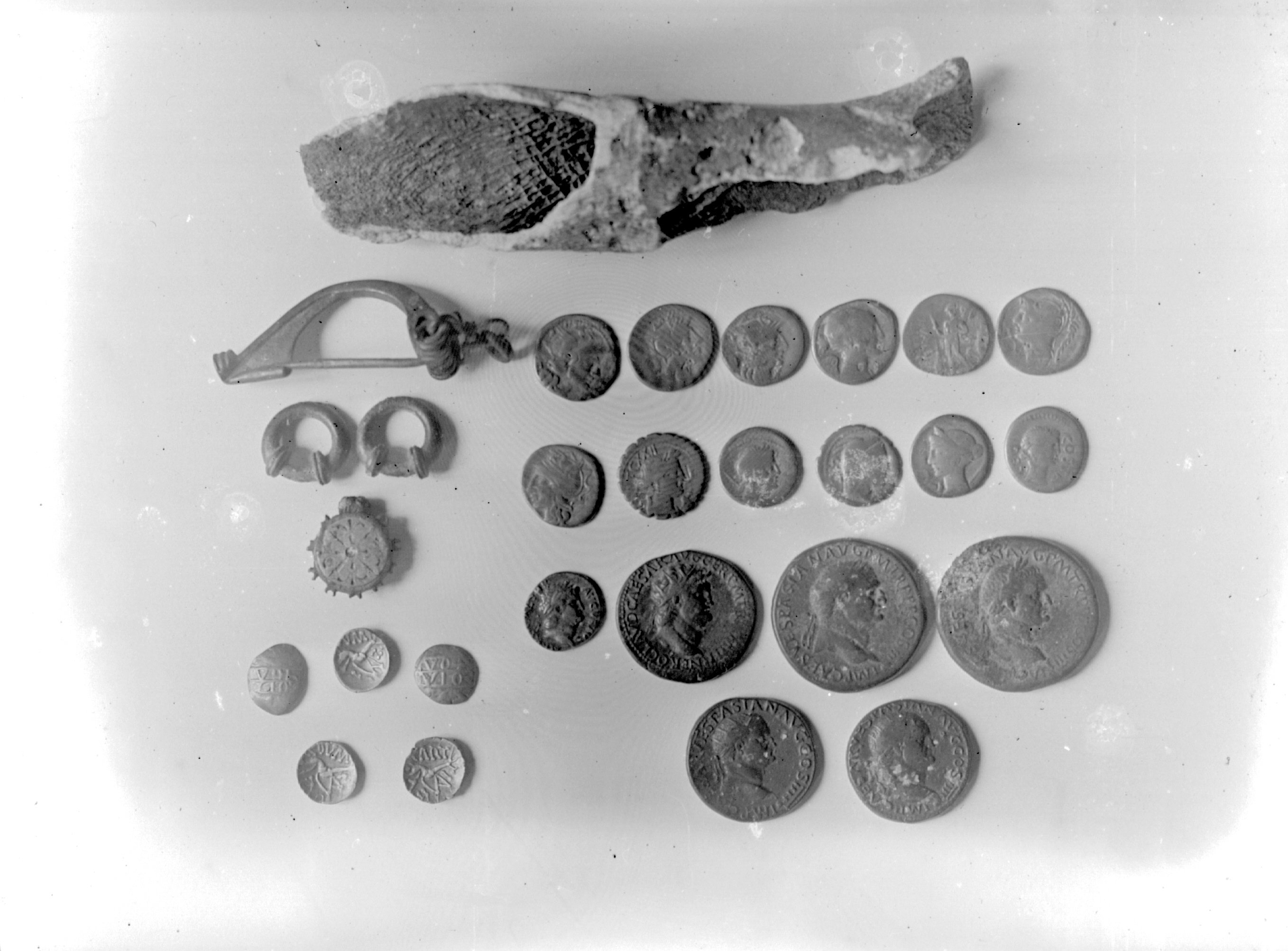 Black and white photograph taken in 1923 showing a collection of Roman artefacts from the Honley Hoard which was discovered in 1893 in Honley, a village in West Yorkshire. The image includes Roman coins, newer silver coins, coins from the Coritani tribe, and Roman trinkets. The items are arranged in neat rows, with detailed inscriptions and profiles of emperors visible on some of the coins. The other trinkets appear to be a fibula (brooch), two earrings, and a decorated wheel-shaped object. At the top of the image lies a piece of pottery or bone with a textured surface. The artefacts are displayed on a plain background for documentation.