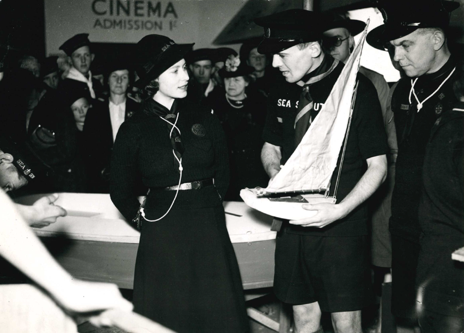 Black and white photograph showing Princess Elizabeth in her Sea Rangers uniform being shown a model boat by Sea Scouts