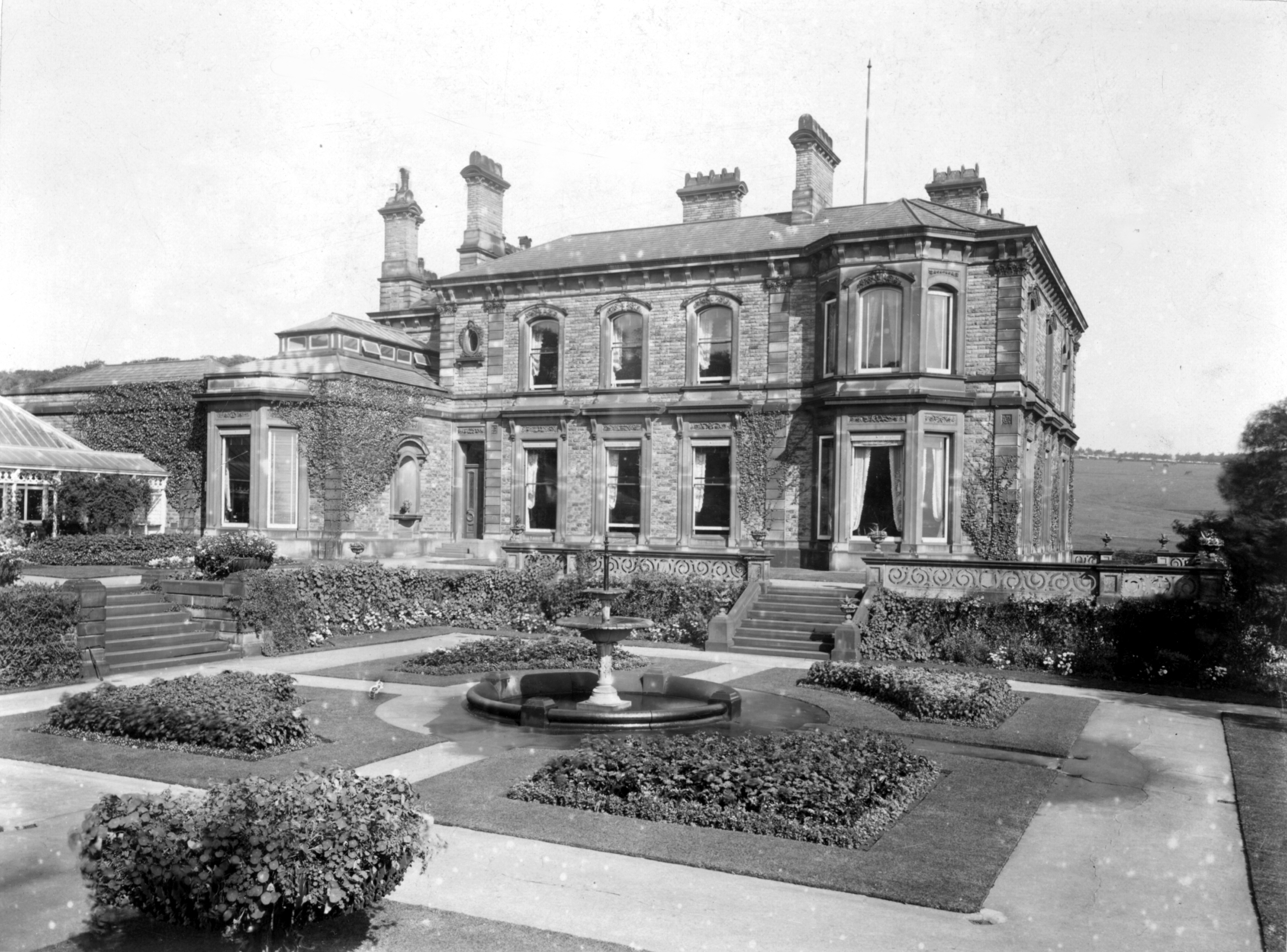 A black and white photograph of a grand Victorian mansion with ivy-covered walls, large windows, and ornate chimneys. In front, a garden features a central fountain and manicured hedges.