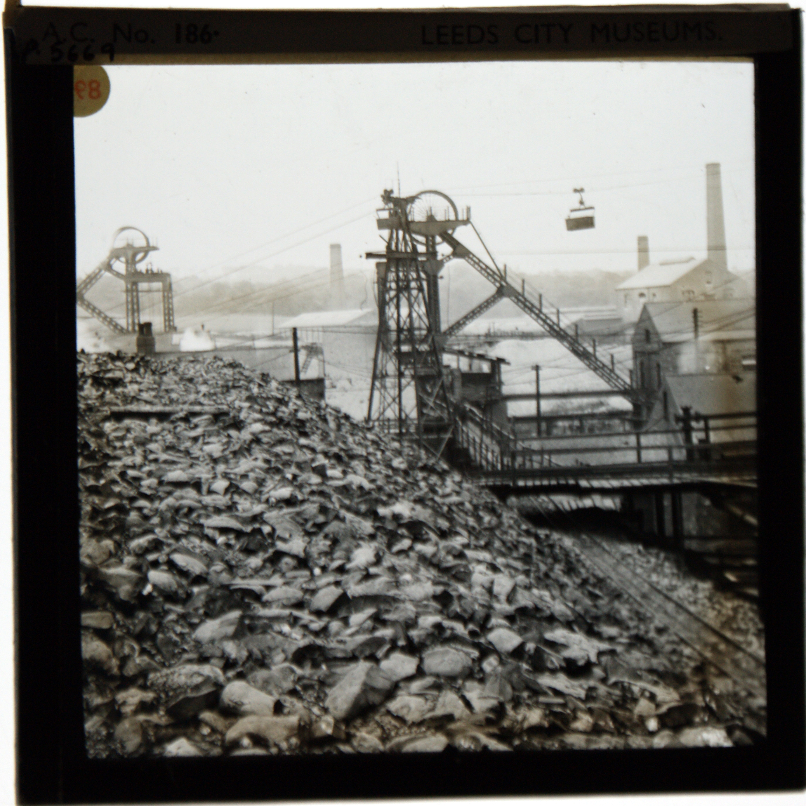 Black and white photograph taken standing on a slag heap, with much stone in the foreground. The winding gear of the colliery is in the middle of the picture, with buildings to the right and behind.
