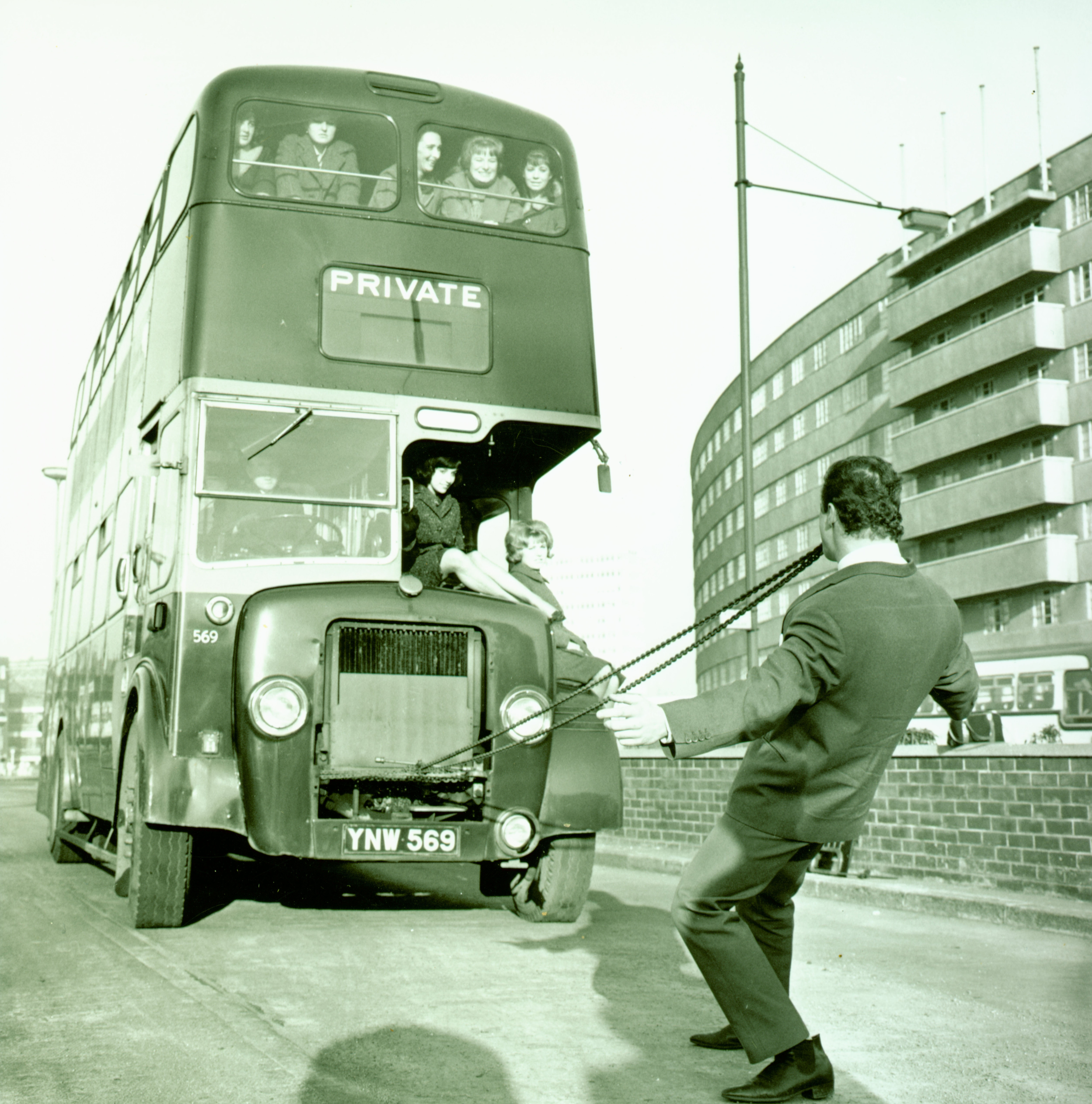 A man is pulling a double-decker bus along a street in Leeds.  One end of the rope is attached to the front of the bus, the other is in his mouth.