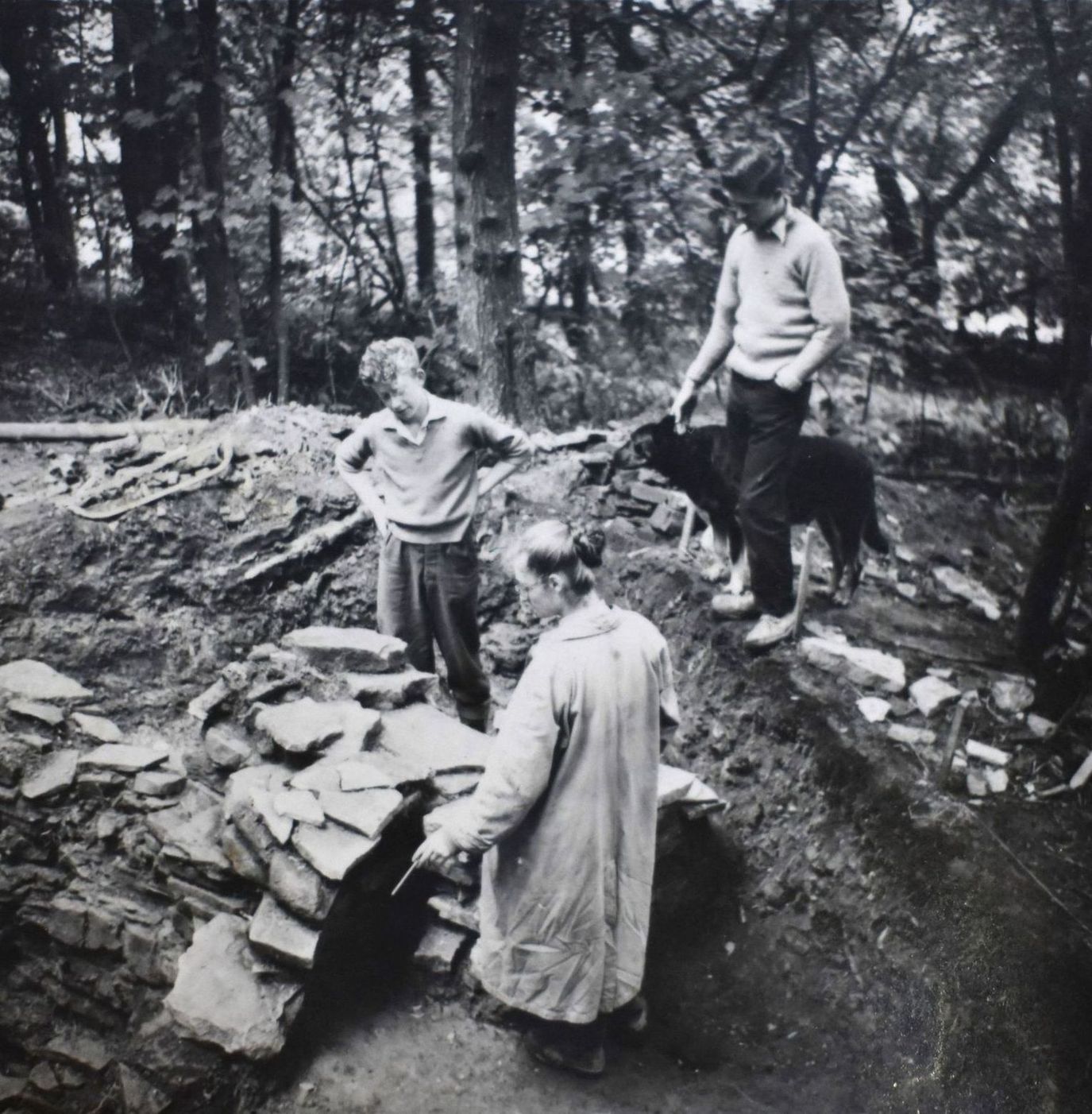 Three people stand at a stone archaeological site in a wooded area. The people are looking into the site, examining it, and one is holding the collar of a large black dog.