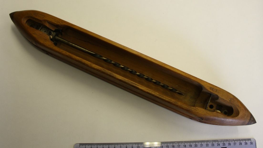 A long wooden weaving shuttle with pointed ends and a metal rod inside, placed on a light surface beside a ruler.