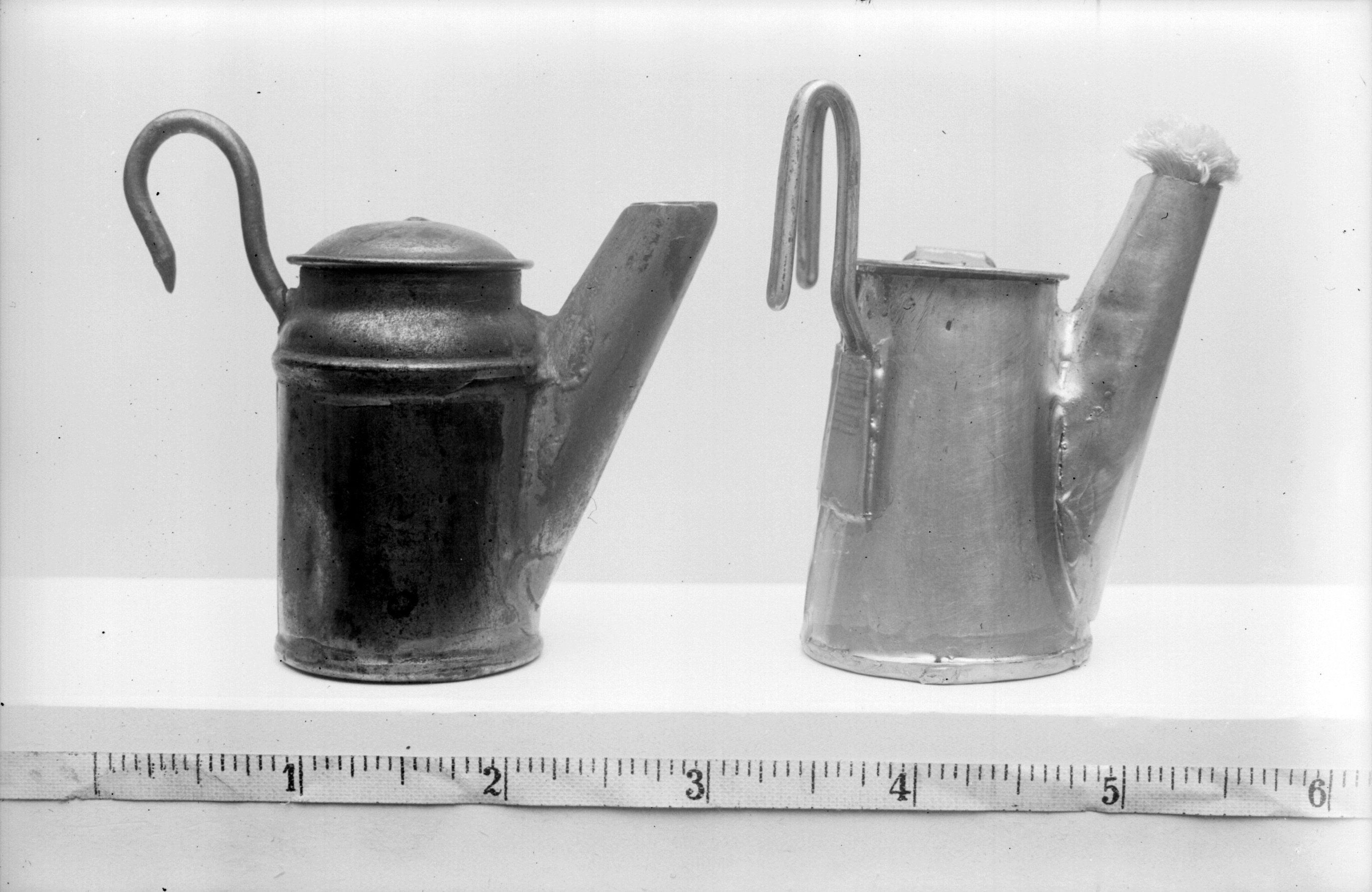 This black and white image shows two small metal miners' lamps placed side by side on a flat surface. Both lamps have a cylindrical body, a spout angled upwards, and a curved hook handle for carrying or hanging. The lamp on the left is darker and has a fitted lid, while the lamp on the right is lighter in colour and features a small brush attached to the end of its spout. A measuring tape below them indicates each lamp is approximately 3 to 4 inches tall.