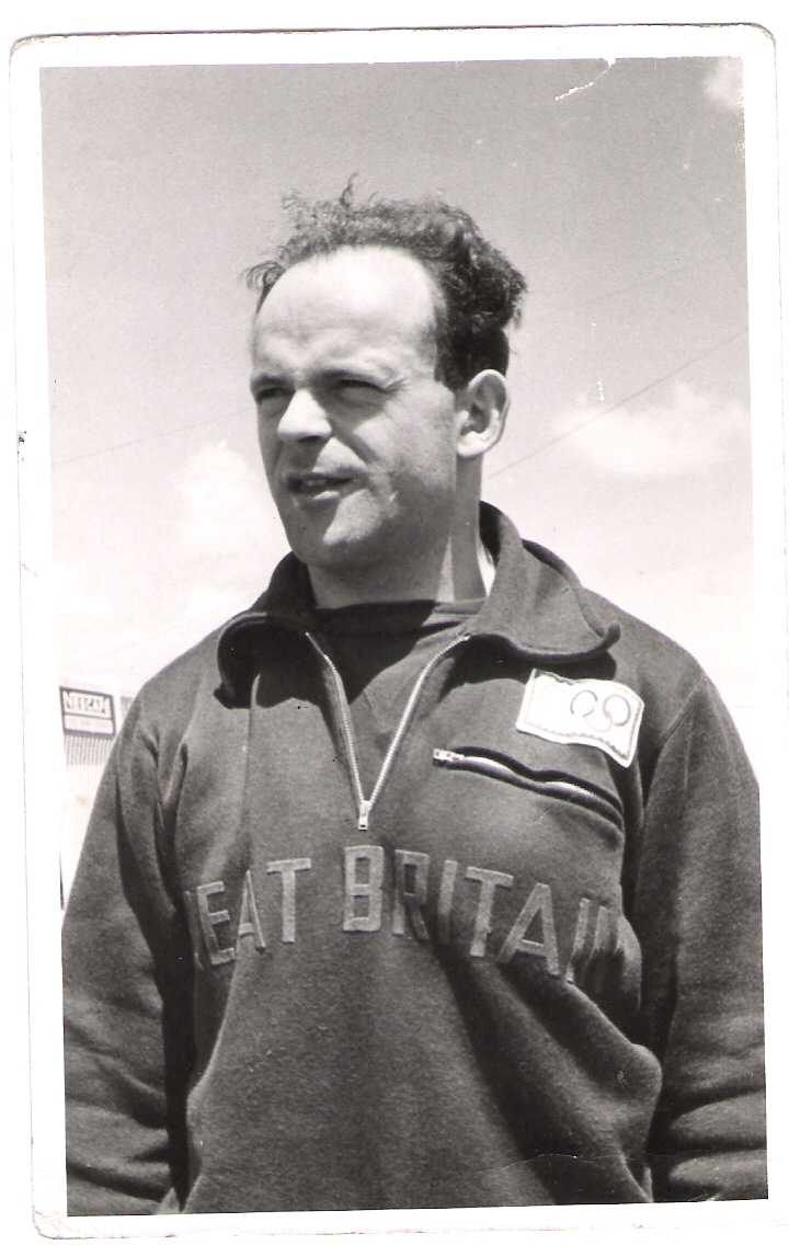 The image is a black-and-white photograph of a Ben Helfgott standing outdoors. He appears to be in his late 30s or early 40s, with short, slightly receding hair. He is wearing a sweatshirt with the words "Great Britain" emblazoned across the chest, along with a patch that includes the Olympic rings symbol.  Taken around the time of the 1956 Olympics.