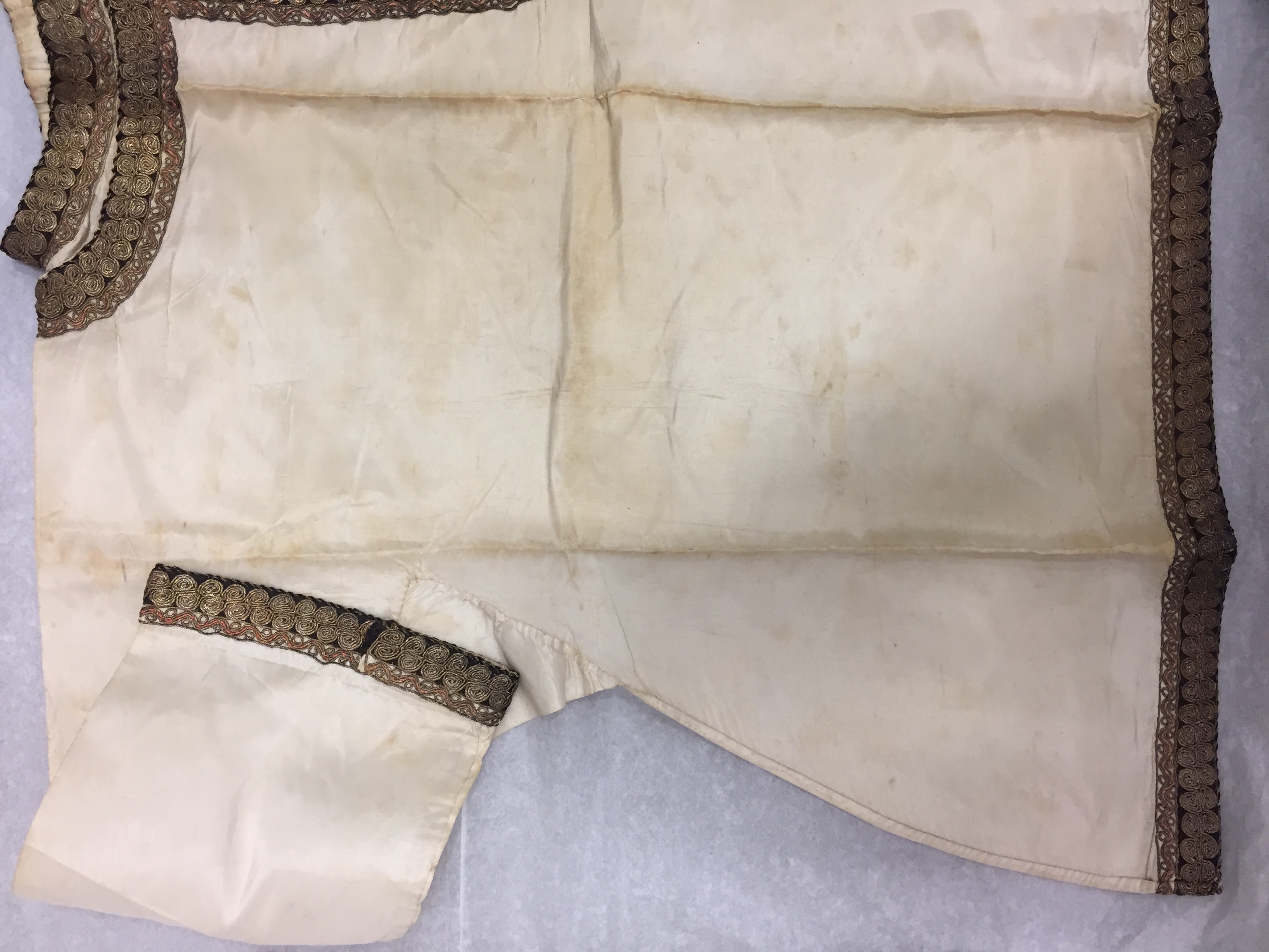 White tunic with brown braid around the cuffs and hem. There are multiple creases across the garment.
