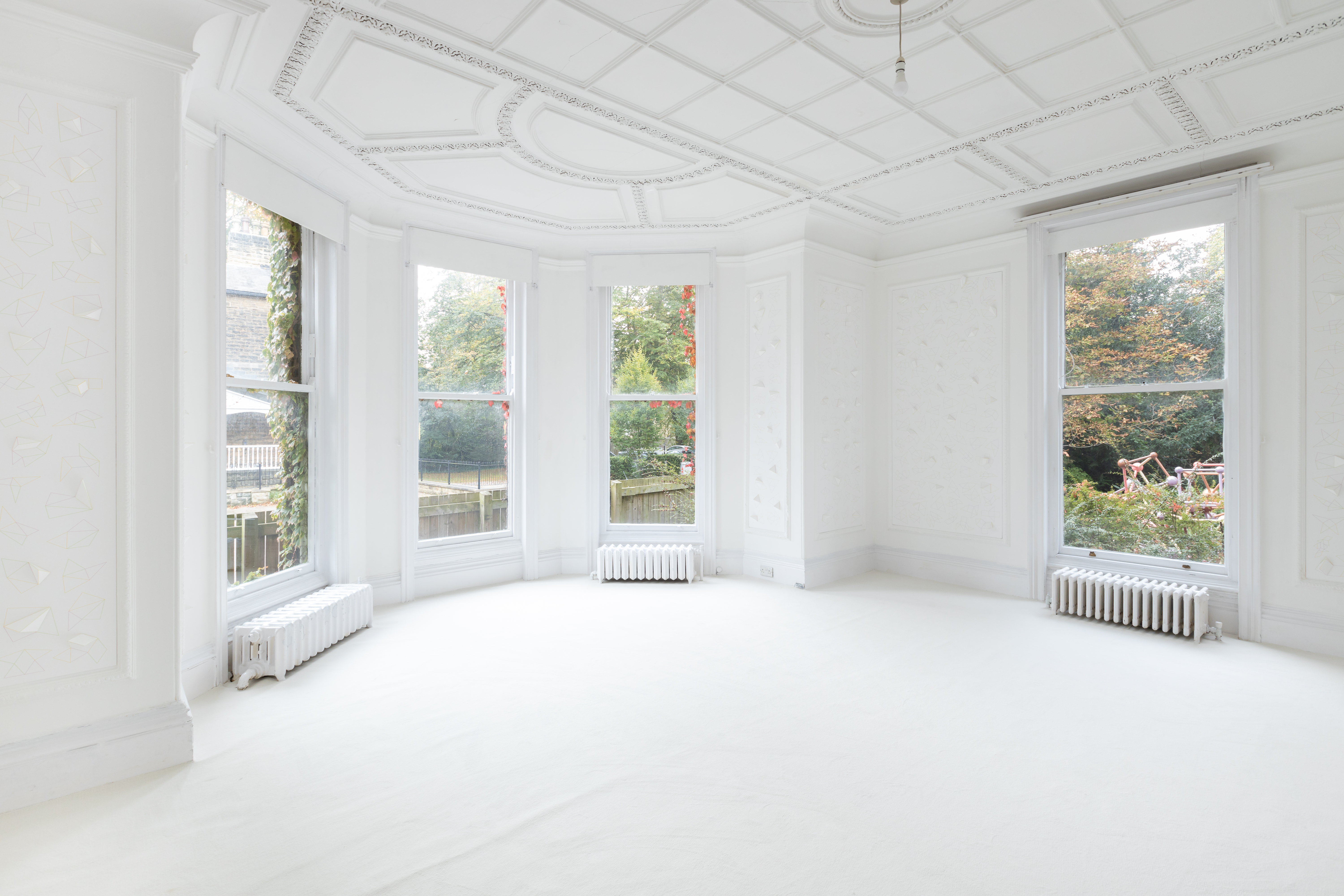 A fully white room with large bay windows overlooking a garden and fence. The ceiling of the window is decorative paneling with coving features. Small metal white radiators are below three of the windows. Through one window there are sculptures of tubes and balls shapes within buses and trees.