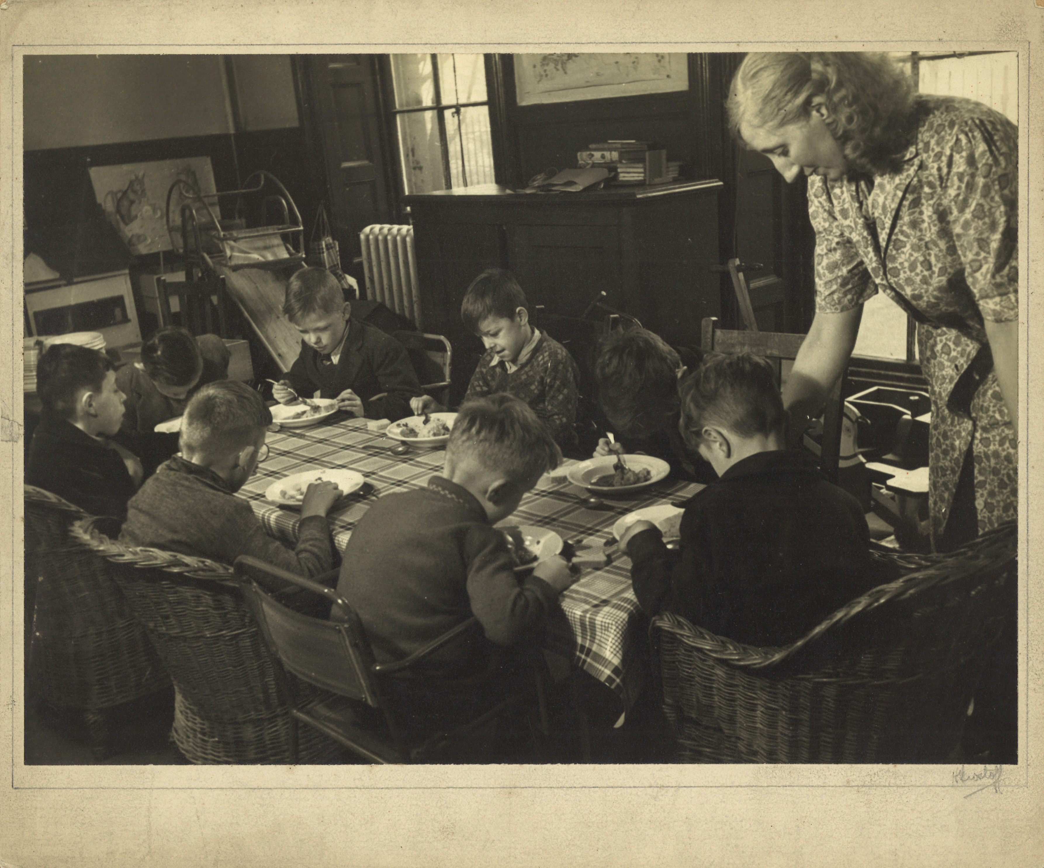 Black and white photograph showing a group of young boys sitting around a table eating.  There is a woman teacher or helper in the picture too.