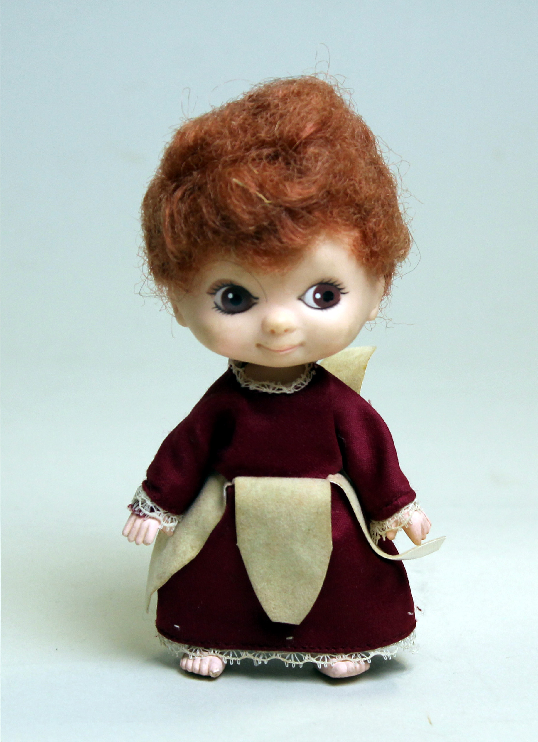Colour photograph of a small doll wearing a dark red dress with a cream petal leaf shaped over-skirt.