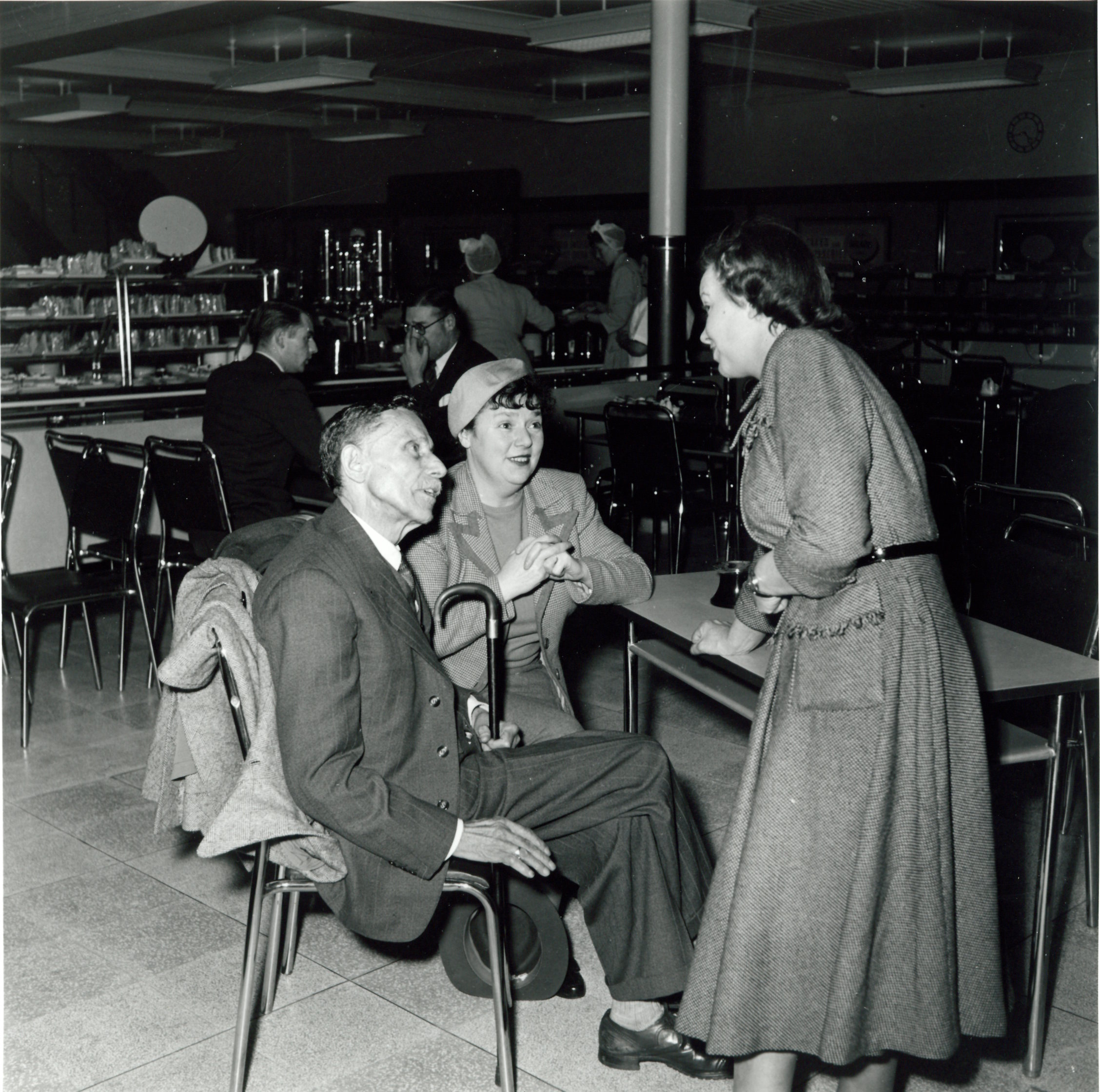 Black-and-white photograph of three adults chatting at a café table inside a mid-20th-century store.