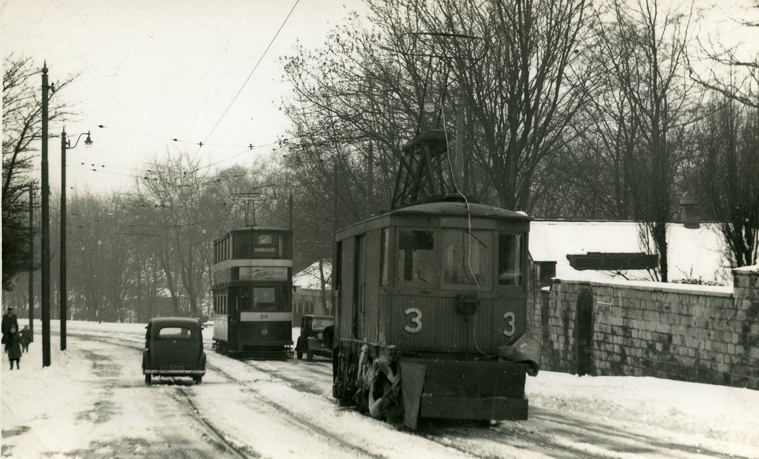 Black and white photo of a road sweeper and trolley bus on a road in the snow coming towards the camera on the right hand side of the picture. On the left is a car going away from the camera. On the far left is a family walking on the pavement