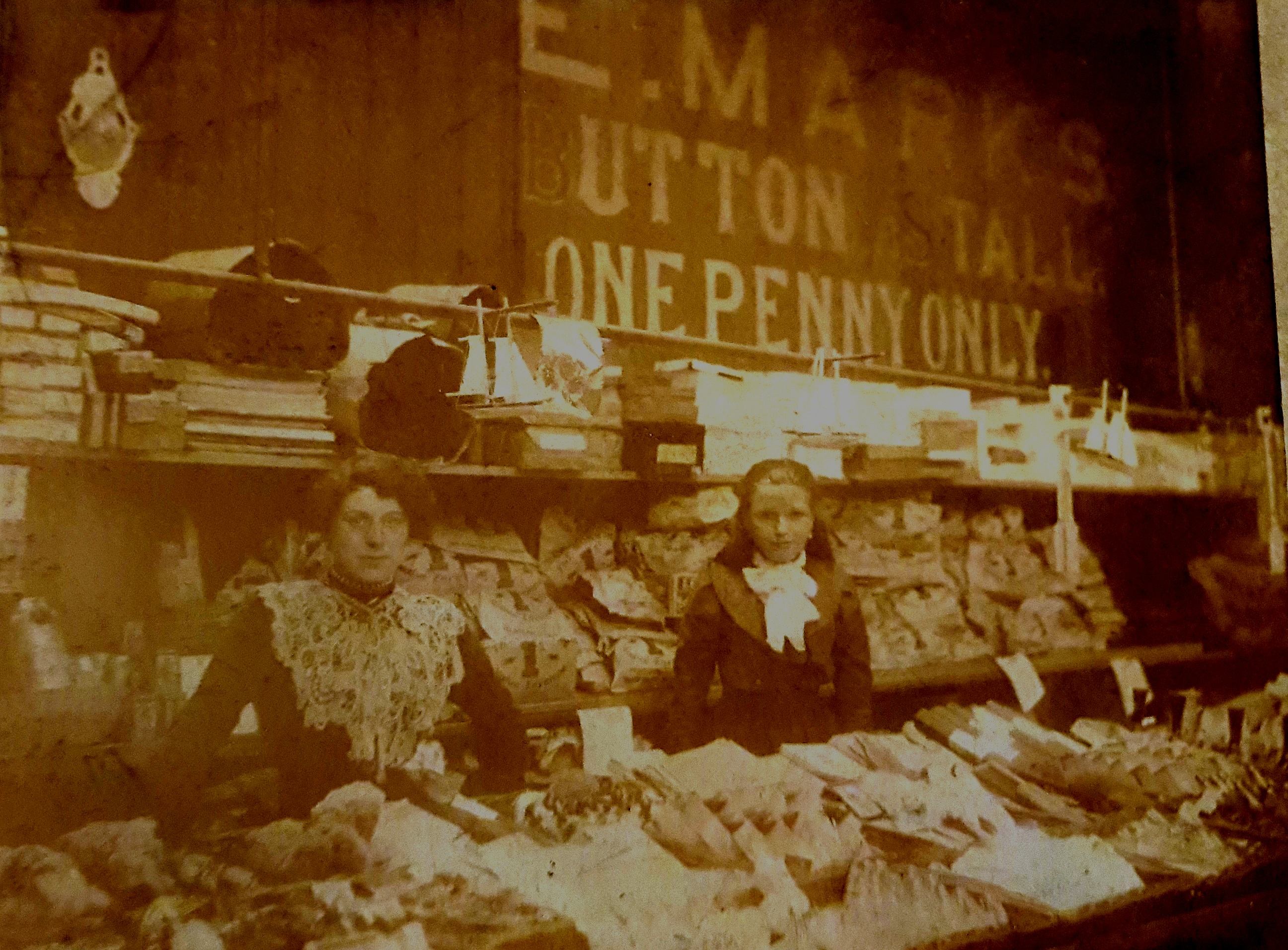 Sepia photo of a market stall with female staff behind the counter and signage E.Marks Button Stall One Penny Only