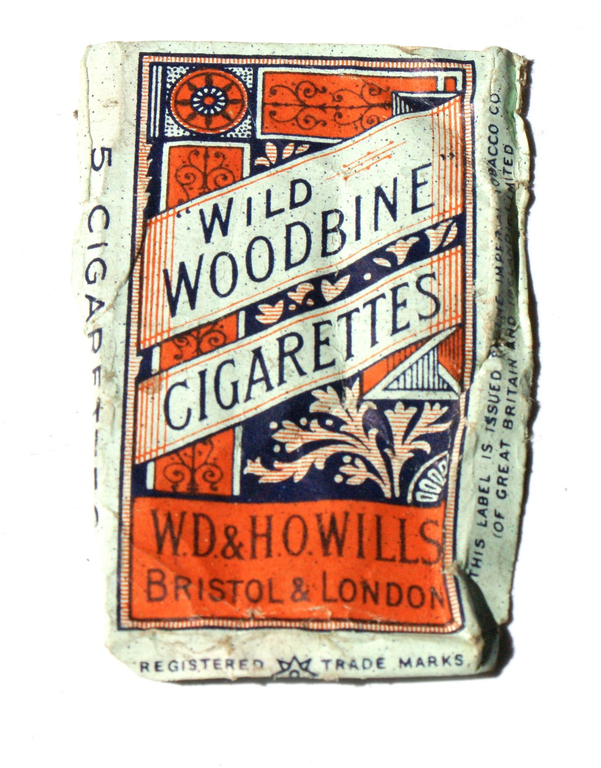 Colour photograph of a packet of cigarettes. It says 'wild woodbine cigarettes' in dark blue text on a design in orange.