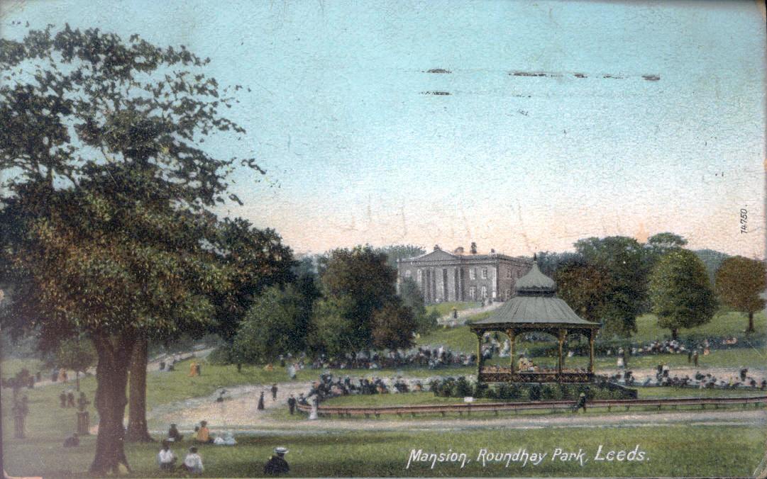 A coloured postcard of a park. On the lef,t is a large tree in the foreground;, on the right in the middle distance is a bandstand, then some trees, and in the far background in the middle is a large Palladian house. Across the picture there are many small figures of people having a day out at the park