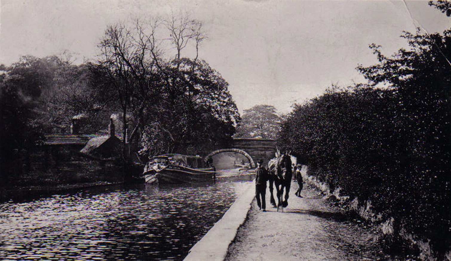 A black and white image of a horse and man walking down a canal path with a canal boat on the water and bridge behind. Houses and smoke coming out of small chimneys can be seen on the left of the image. Trees can be seen on both side of the image