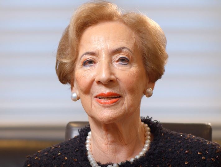 The image is a color photograph of an elderly woman with short, neatly styled hair. She is wearing pearl earrings and a pearl necklace, complementing her elegant attire, which appears to be a dark, textured jacket. Her makeup includes bright red lipstick, and she has a warm expression, smiling gently at the camera. The background is softly blurred, focusing attention on her face, which exudes wisdom and grace.