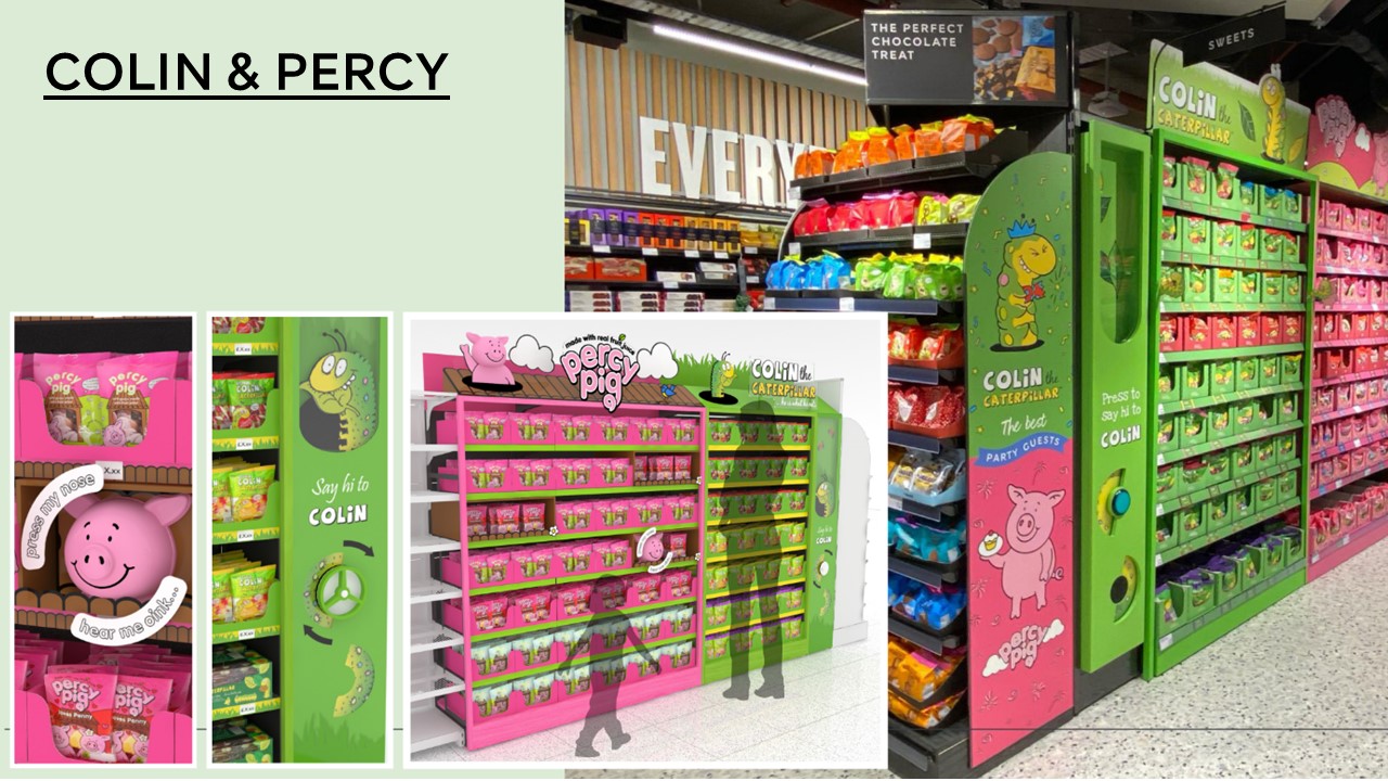 Marks & Spencer supermarket aisle displaying colourful Colin the Caterpillar and Percy Pig product stands. Bright pink and green packaging with cartoon characters creates a playful atmosphere. Shelves are filled with various sweets and chocolate packets.