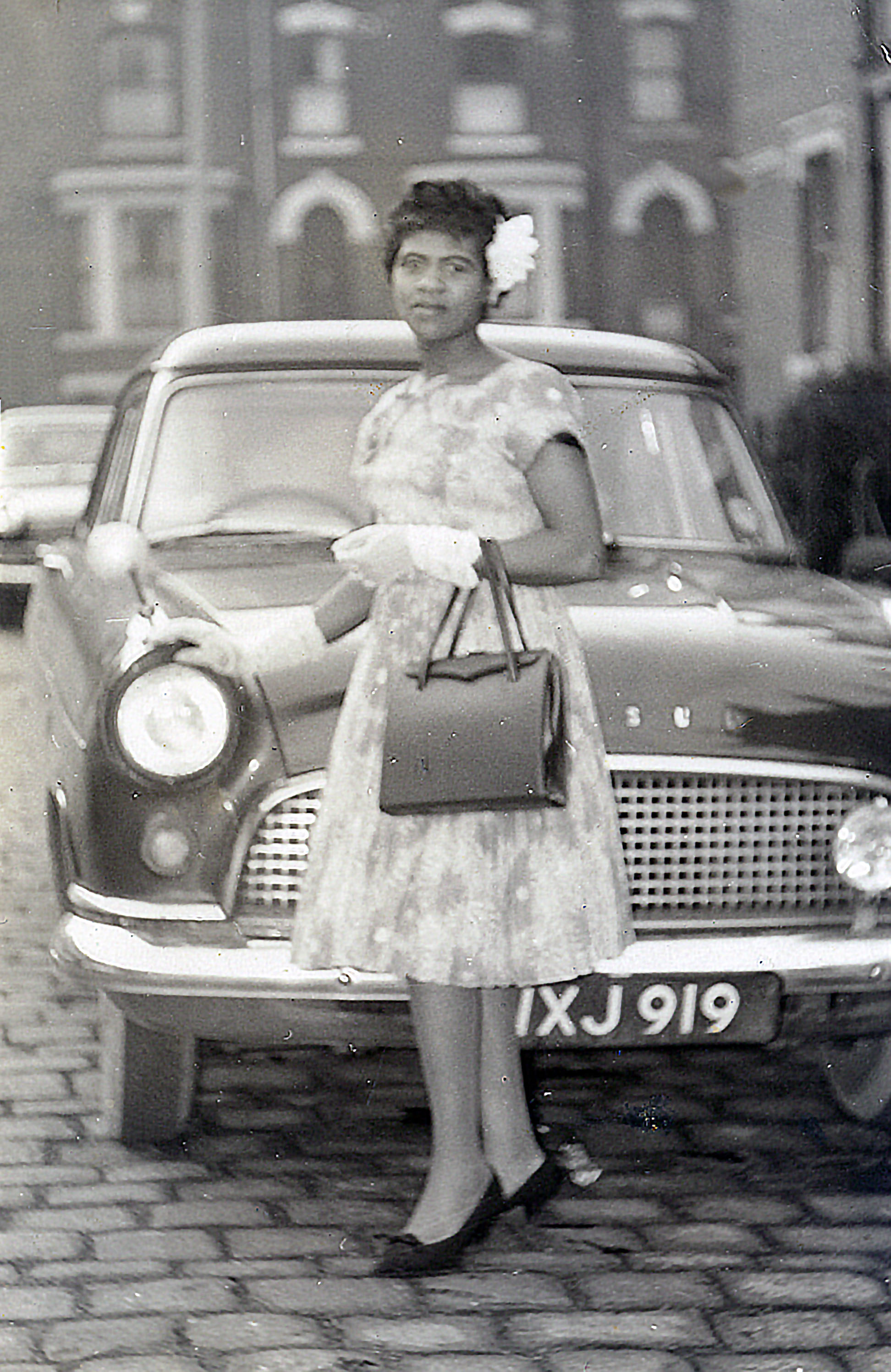 Black and white photograph showing a young woman in a white dress and gloves, standing in front of a car.