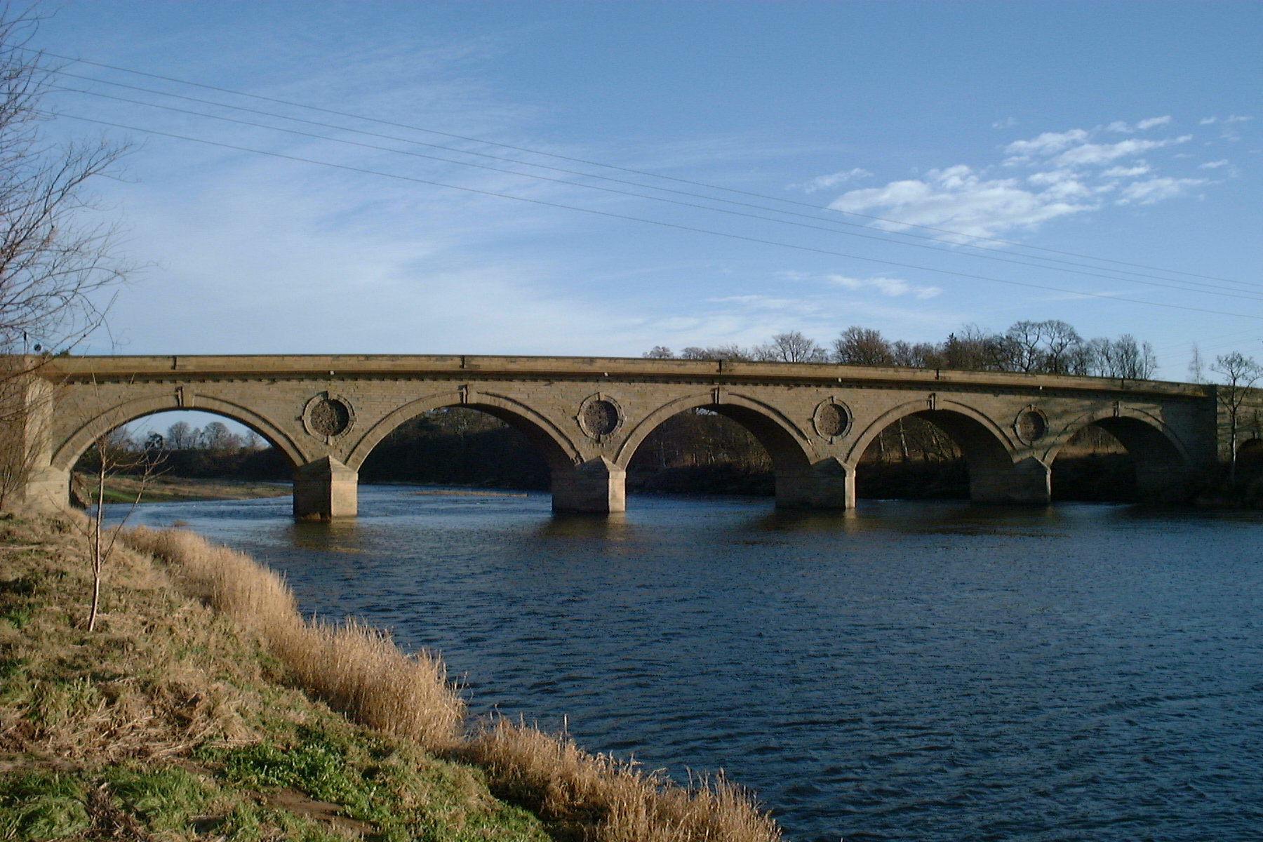 Light blue sky with light clouds. Stone built bridge running across the centre of the image. AT the bottom of the image is a grass bank and dark blue water