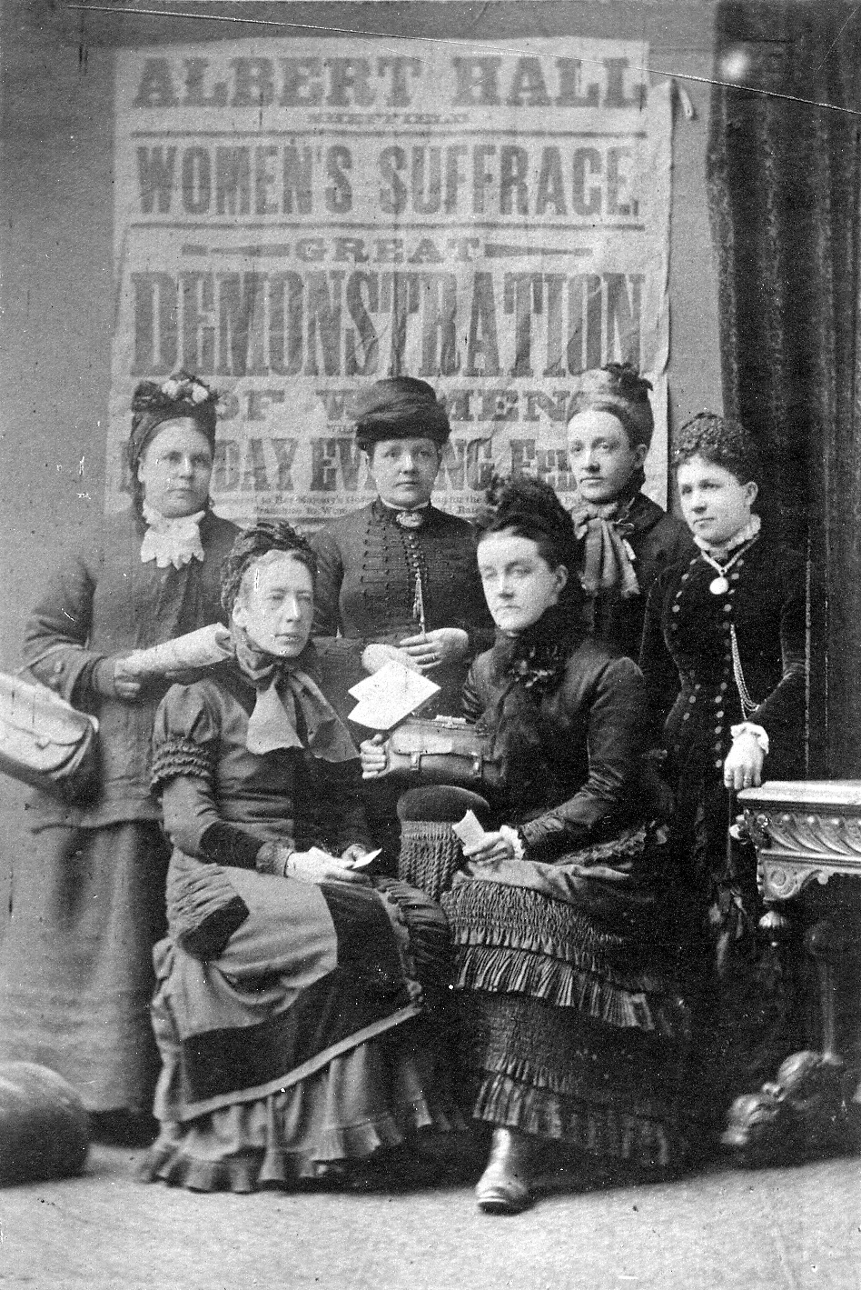 Black and white photograph showing a group of women in Victorian period dress in front of a Women’s Suffrage demonstration poster