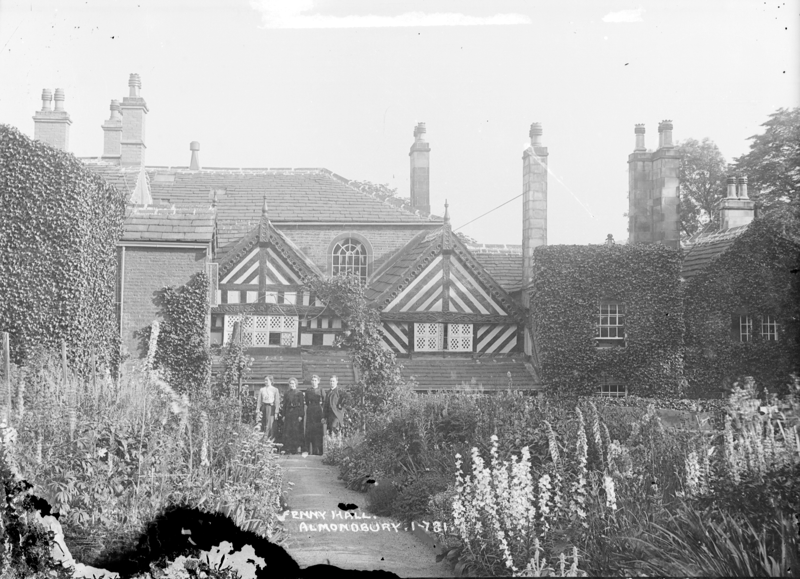 A historic, ivy-covered brick house with striped gables and tall chimneys. Five people stand in front, surrounded by tall wildflowers. White text at the bottom of the image labels it as Fenay Hall, Almondbury.