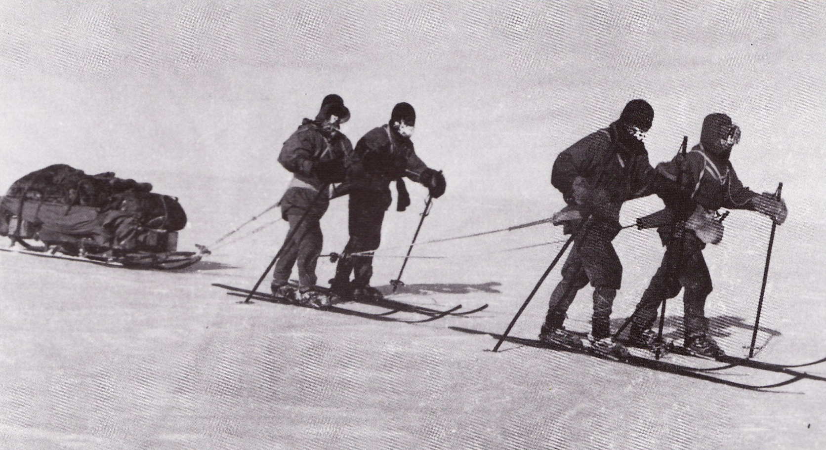 Black and white photograph showing four men on skis pulling a sled laden with supplies