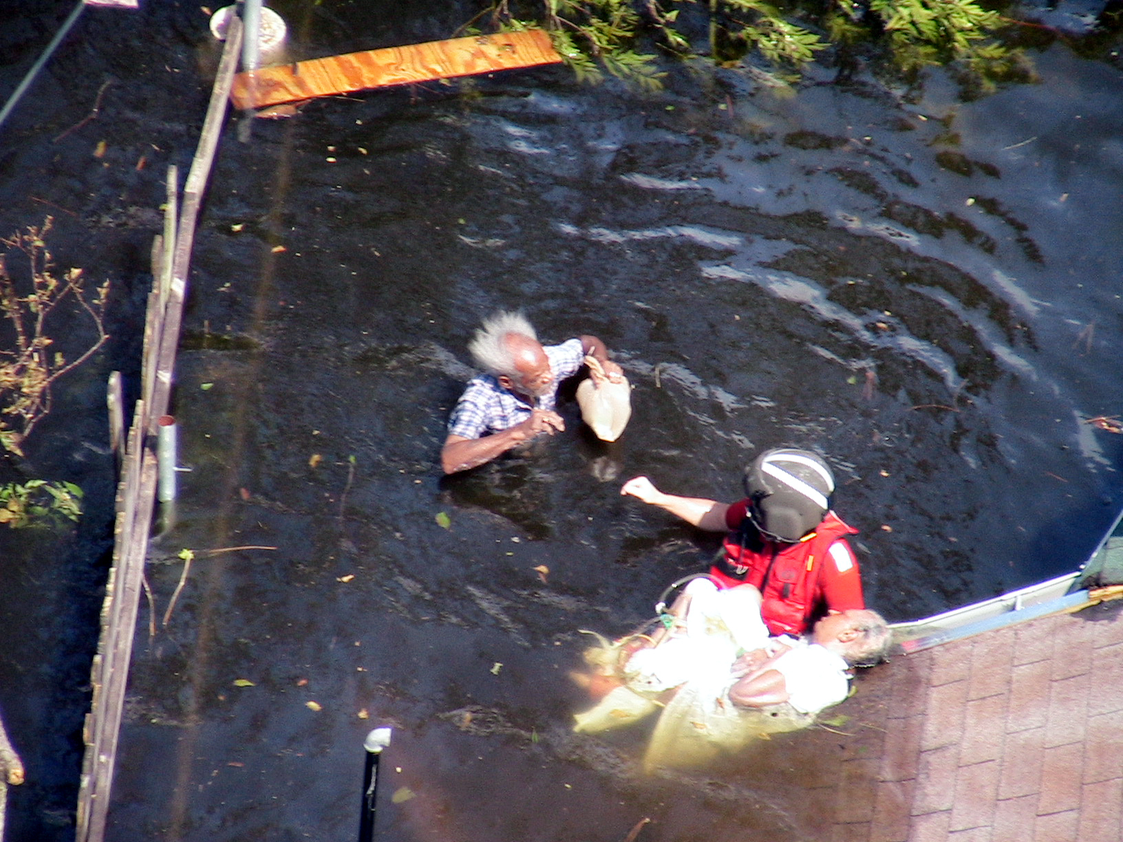 A coastguard wearing a life vest and helmet is in the water with an elderly man and woman.  The woman his lying on a roof, with most of her body in the water.