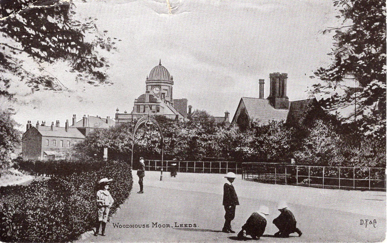 Black and white photograph showing a wide path going ahead and turning slight left. There is a hedge on the left hand side of the path, houses and municipal building, one with a cupola, in the background and children playing on the floor in the foreground.