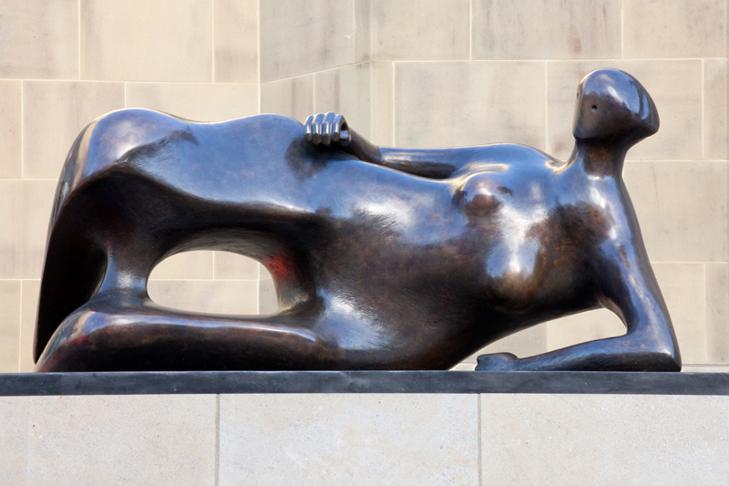 Reclining Woman by Henry Moore, abstract bronze sculpture of a woman lying on her left side, propped up on elbow, with her right knee raised. Her right arms rests on her right side. She looks down along her body