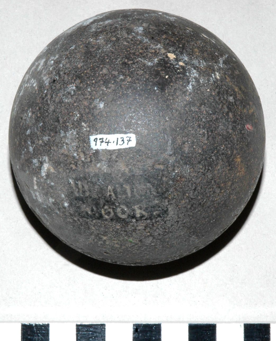 A weathered black cannonball with a small white label "974.137" sits on a light grey background. Below it is a black and white scale for size reference.