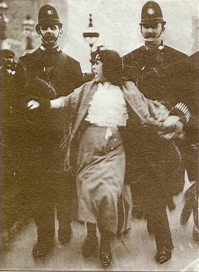 Black and white, grainy, historical photo of a woman in a long skirt and ruffled blouse being escorted by two British police officers in traditional uniforms. She appears to be resisting as they hold her arms.