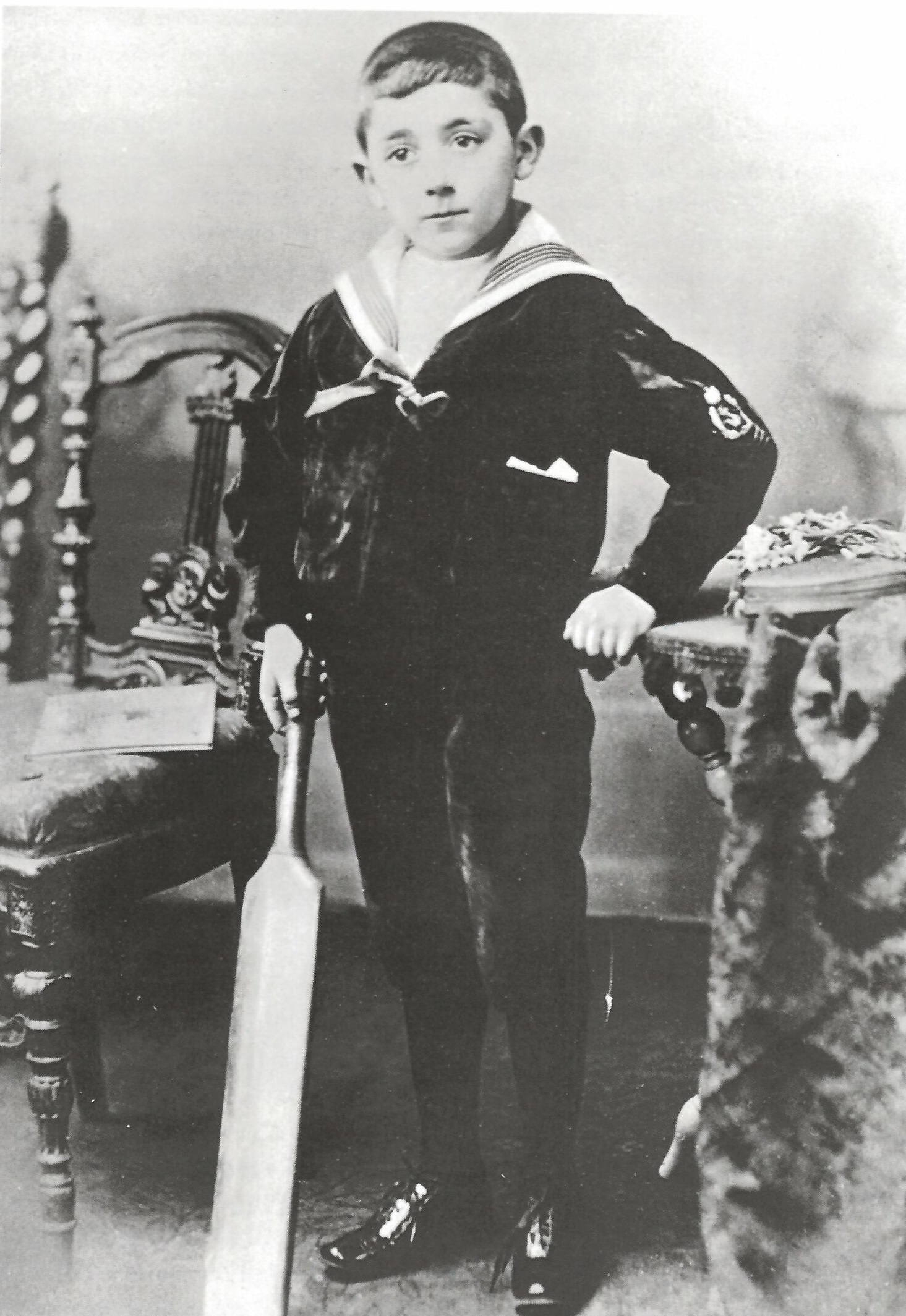 Black and white photo of young boy in sailor suit next to a table and chair holding a cricket bat