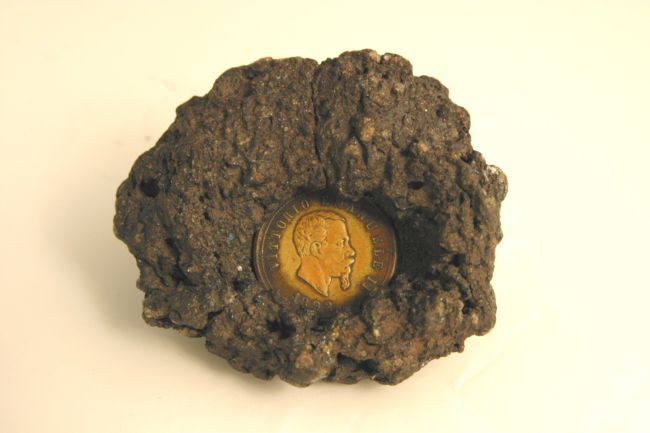 Gold coloured coin stuck in a piece of dark coloured rock.  The edges of the rock curl over the coin, trapping it within the rock.