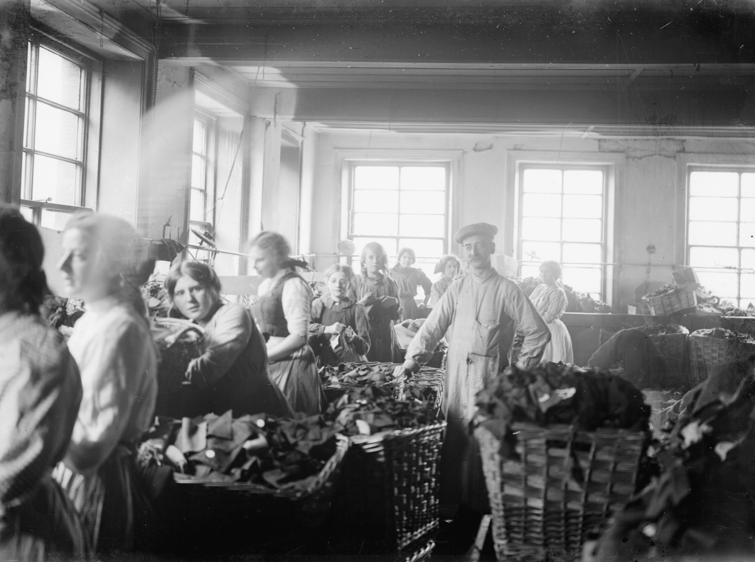 Black and white photograph showing a group of women and one man working in a factory. They are sorting and stripping Army uniforms for buttons and rags. Large baskets filled with fabric scraps and uniform pieces surround them. The women wear aprons and simple dresses, and the man wears a work coat and cap. The room is well-lit by large windows in the background. The scene captures a busy and industrious atmosphere, in the early 20th century.