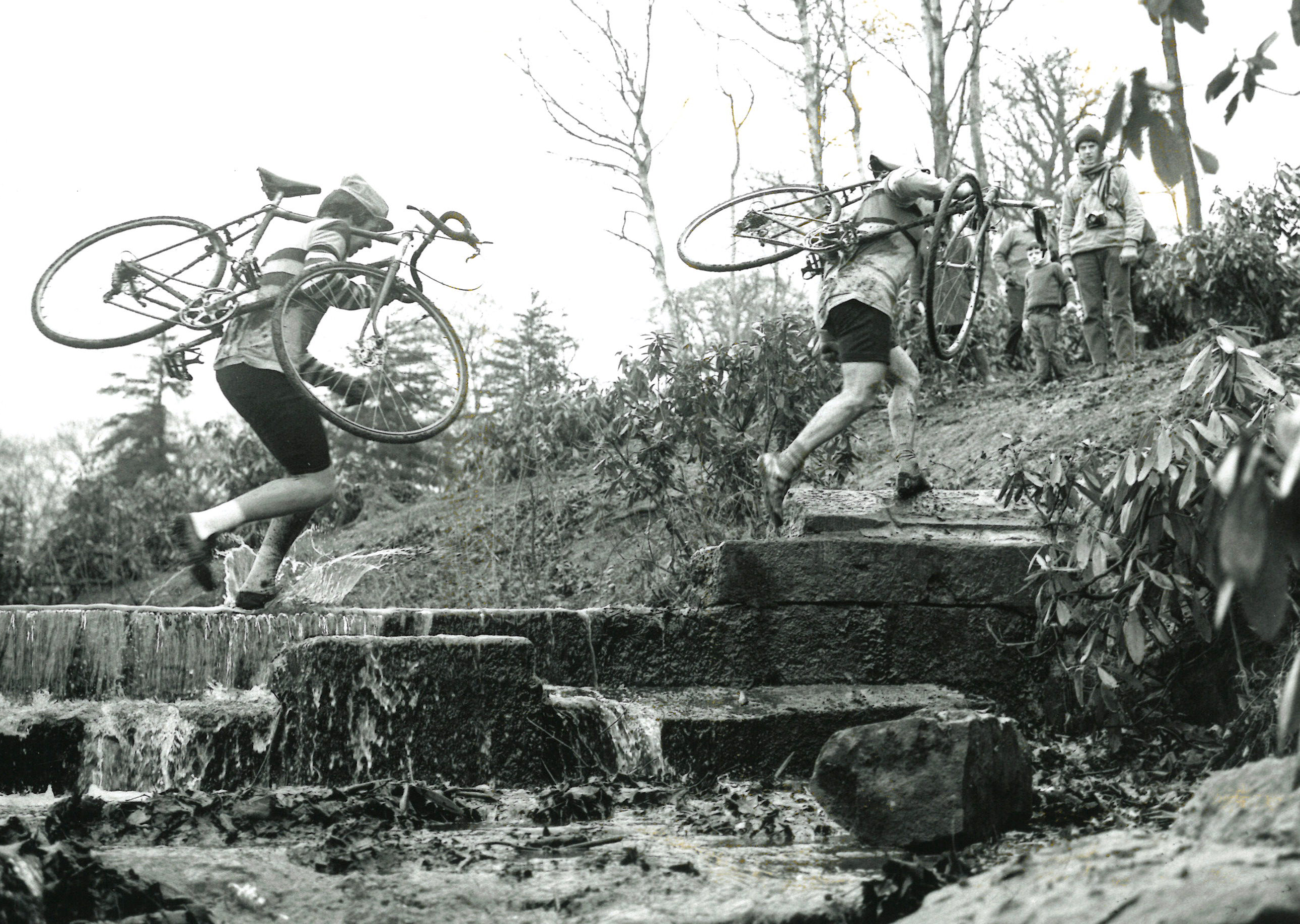 Black and white photograph showing two competitors carrying their bikes over a small bridge