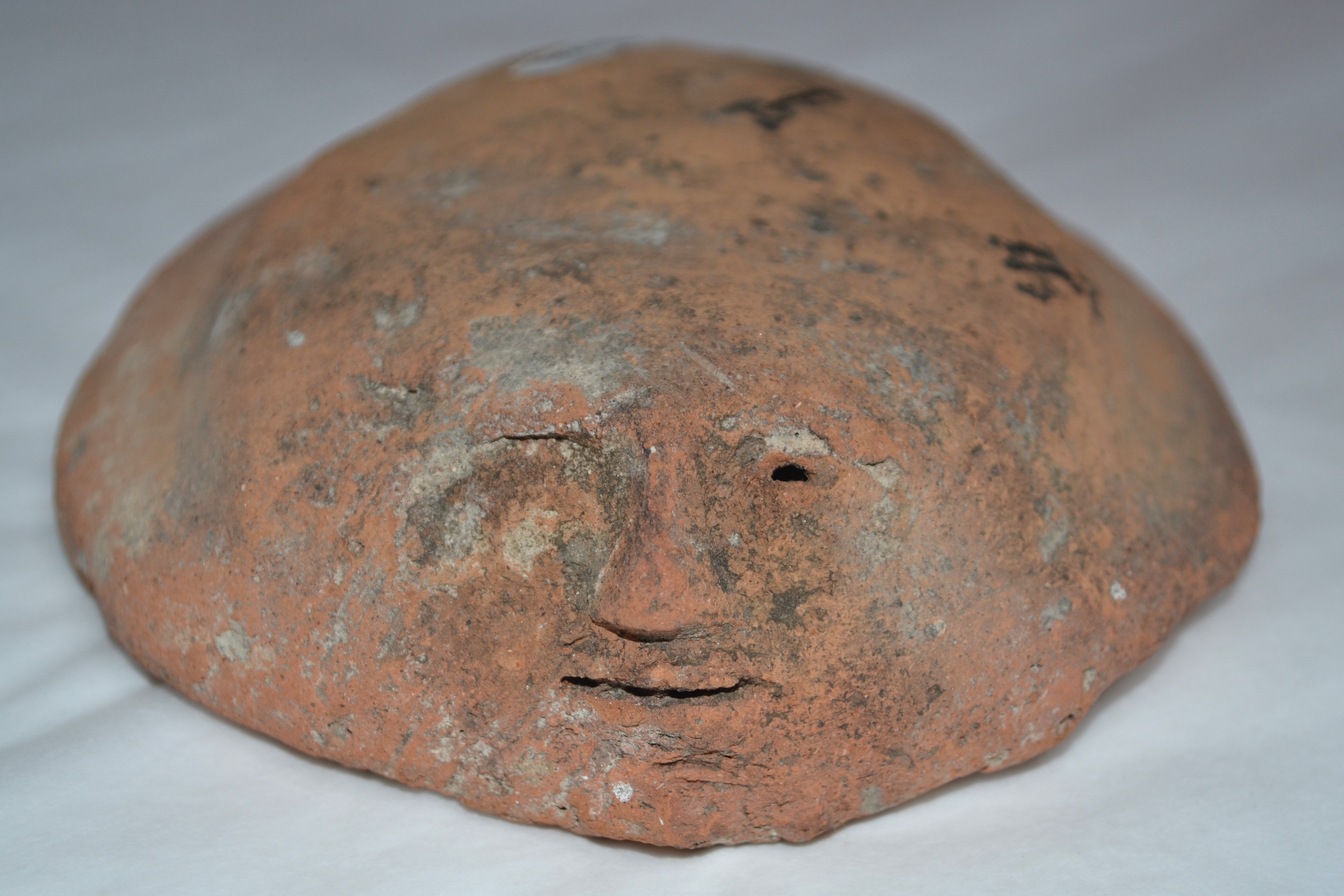 A weathered, orange-brown canopic jar lid shaped like a human face. The facial features are simple and worn, with visible eyes, a nose, and a slightly curved mouth. The surface appears rough, showing signs of age and erosion. A small scale in the foreground indicates the lid’s size, suggesting it is a relatively small artefact.