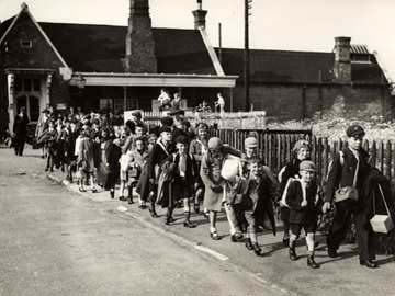 Black and white photo of a line of children walking along a pavement by the side of a fence by a railway track.