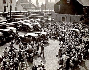 Photograph of evacuees congregating outside in Nottingham.  They are in what looks like a car park, with a line of double decker buses on the road outside.