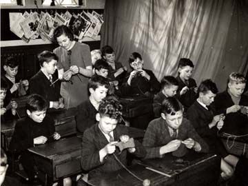 Photograph of Gotham schoolboys knitting at their desks.