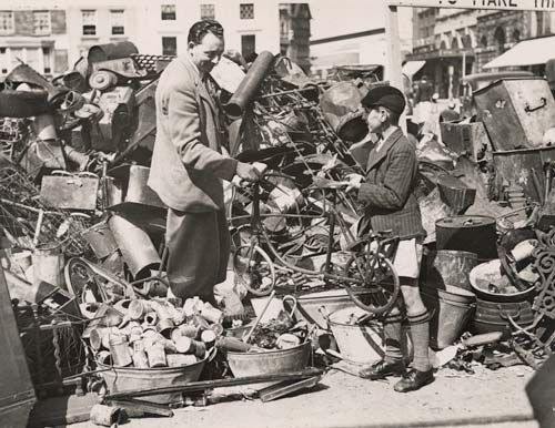 Black and white photograph of a man and a boy surrounded by piles of metal things such as cans, bicycles, pans etc