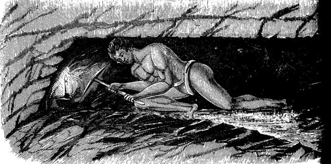 a man is hunched over in a semi lying position in a narrow tunnel.  He has a candle and a pickaxe and is working at the coalface.