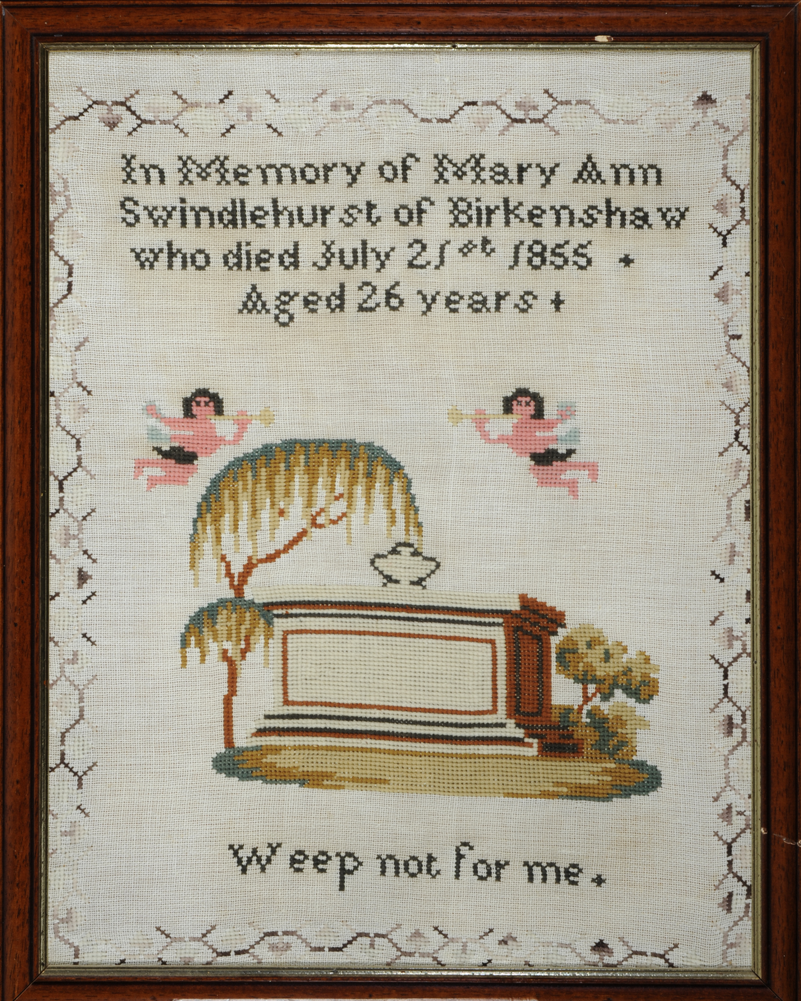 A rectangular wooden frame with gold trim presents a cross-stitch sampler. The sampler has a patterned border and text reading 'In memory of Mary Ann Swindlehurst of Birkenshaw who died July 21st 1855. Aged 26 years. Weep not for me'. There is an image of a coffin surrounded by trees and two angels flying above it.